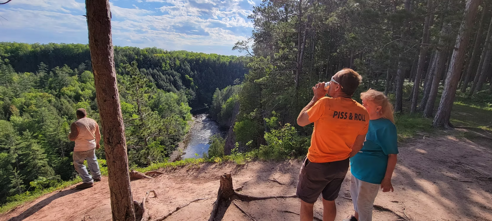 People standing at a forest overlook above a river, with trees and bright sunlight.