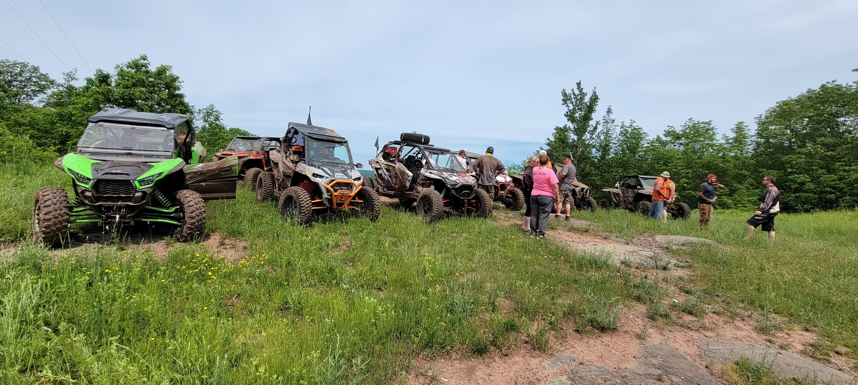 Off-road vehicles lined up on a grassy hill with several people standing nearby under a cloudy sky