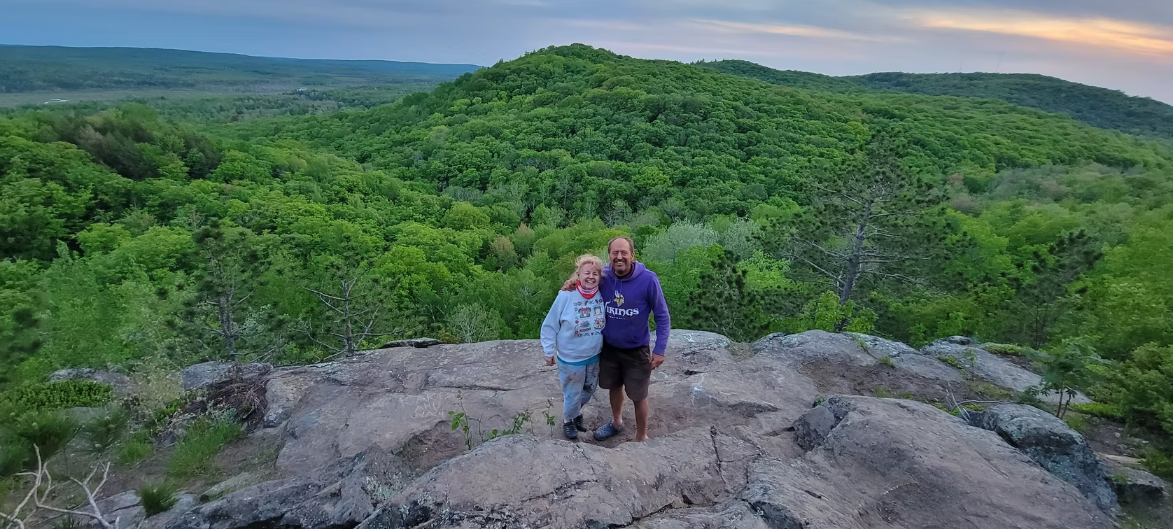 Two hikers standing on a rocky overlook above a lush green forest valley