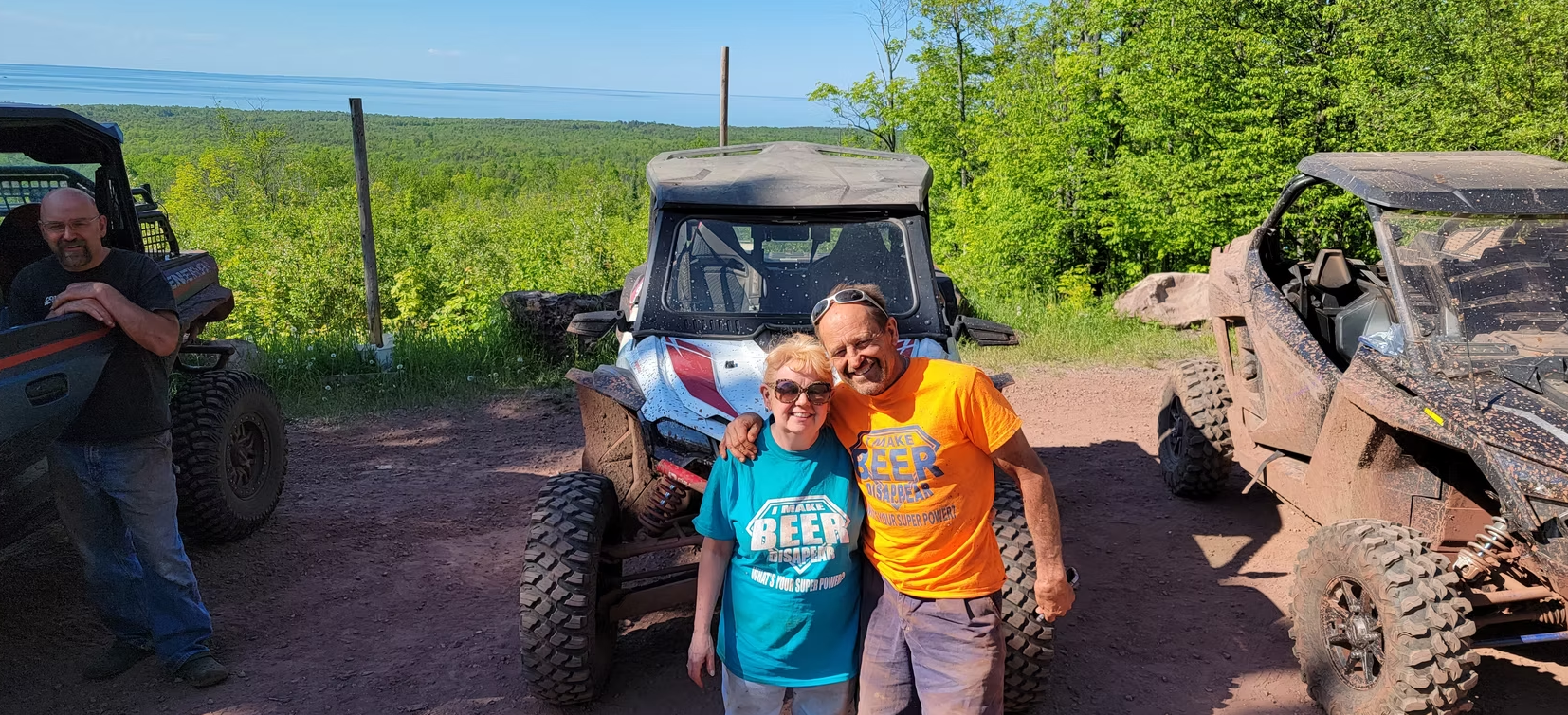 Three people pose smiling in front of off-road vehicles beside a green vineyard on a sunny day