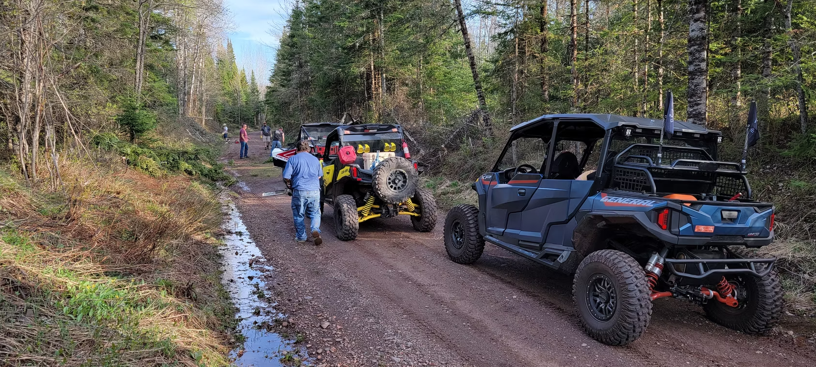 Off-road vehicles and people gathered on a muddy forest trail, with a blue UTV in the foreground