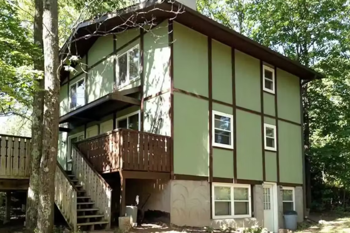 Two-story green house with brown trim, exterior stairs, and a wooded setting