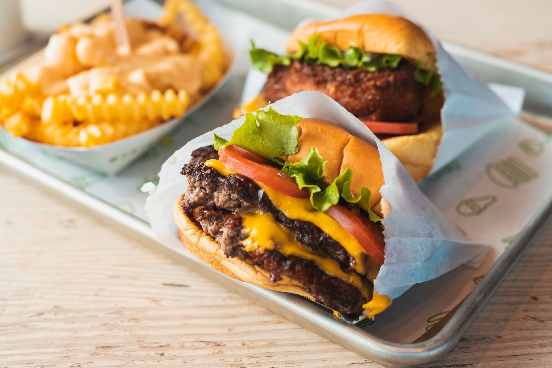 Three burgers and fries on a tray, with one cheeseburger in front topped with lettuce, tomato, and pickles