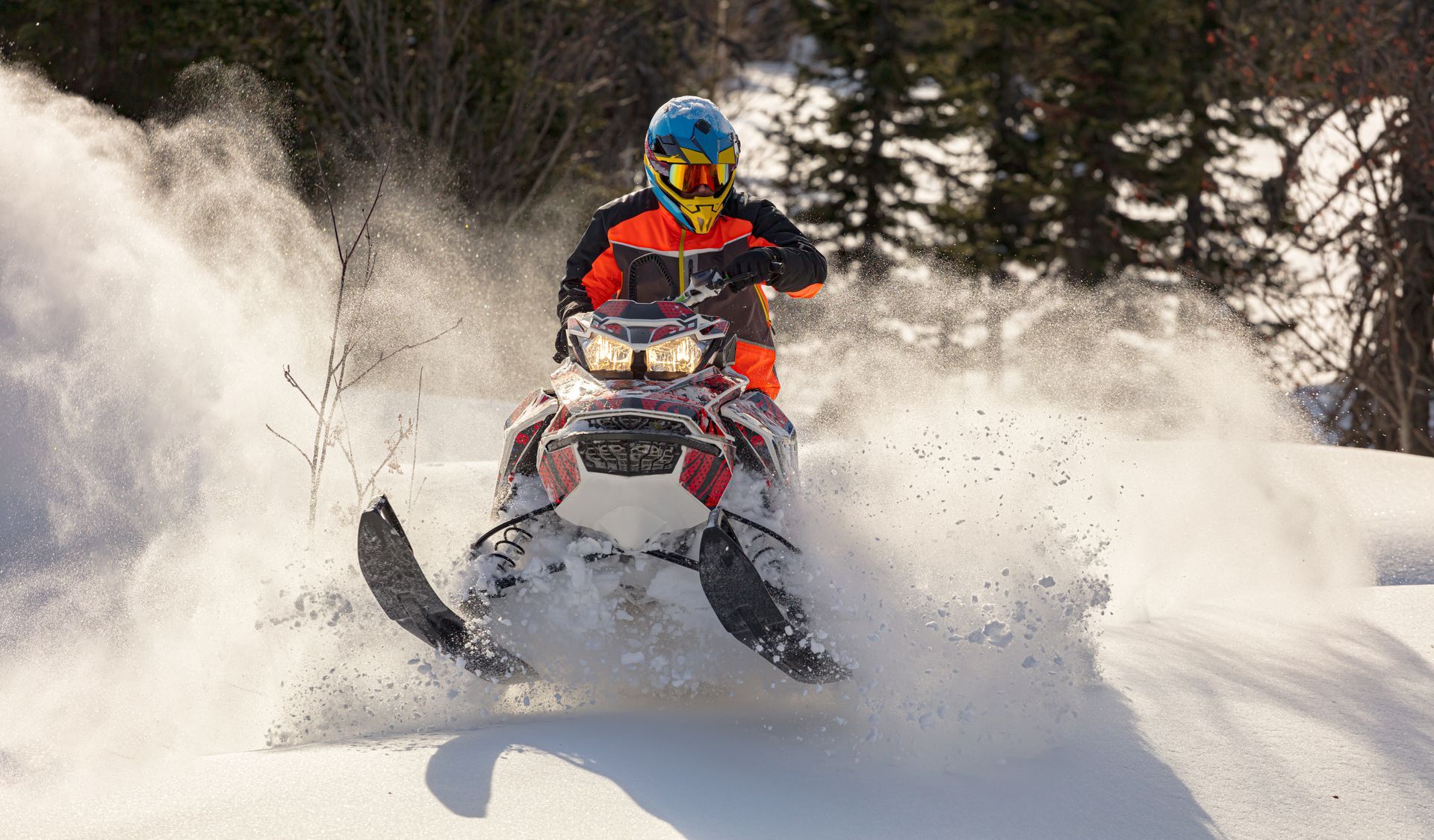Snowmobiler in red gear speeding through deep snow, kicking up powder in a forested winter landscape