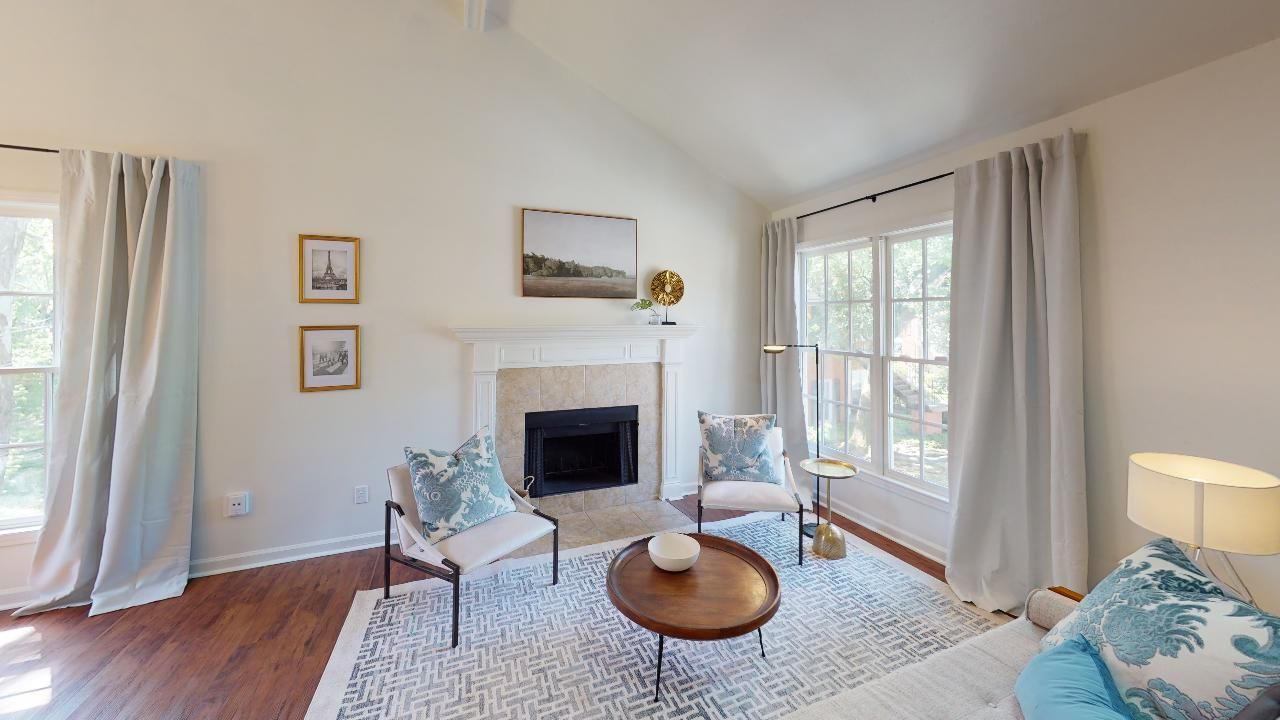 Cozy living room with fireplace, chairs, and coffee table. Cream walls, wood floors, and large windows.