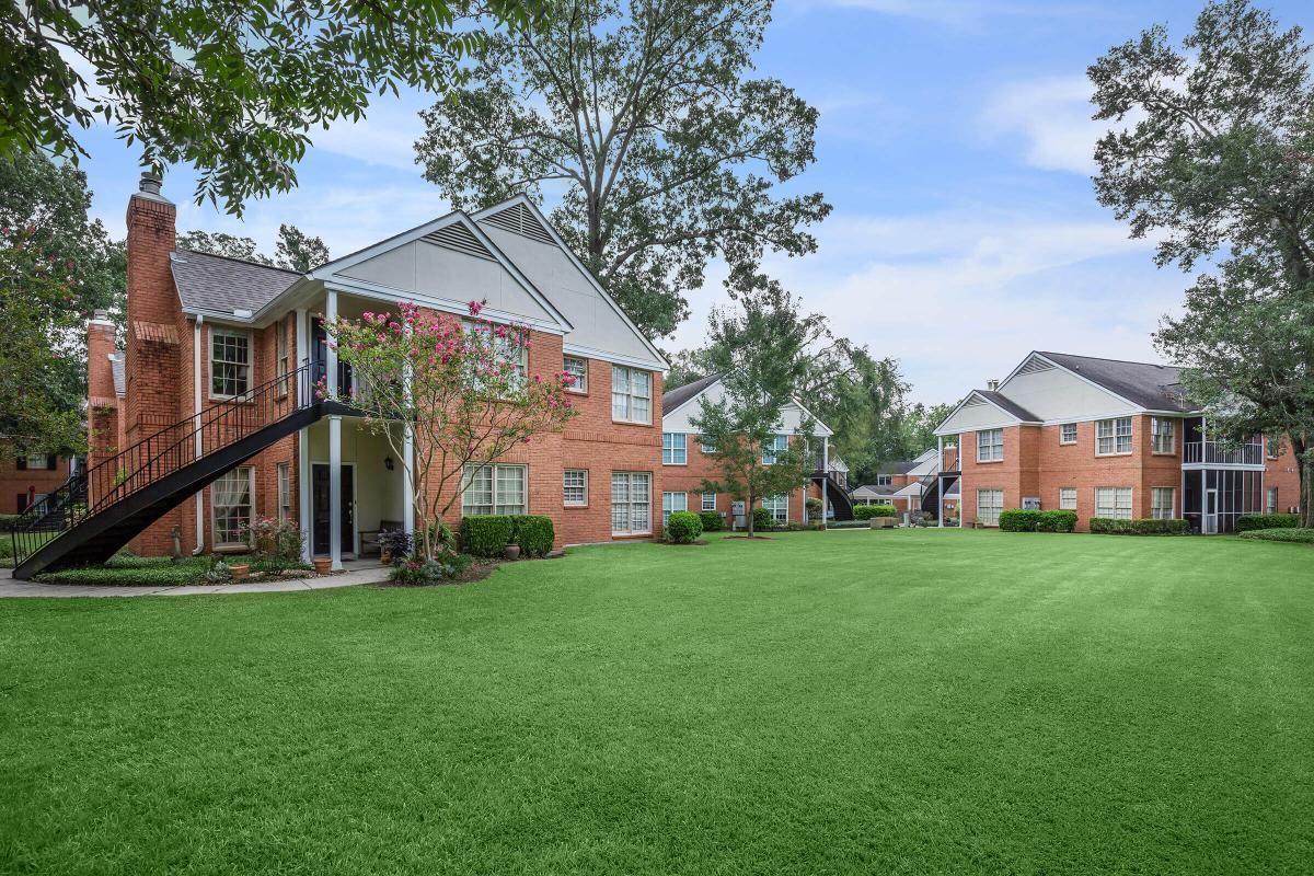 Brick apartment buildings with a green lawn and trees under a cloudy sky.