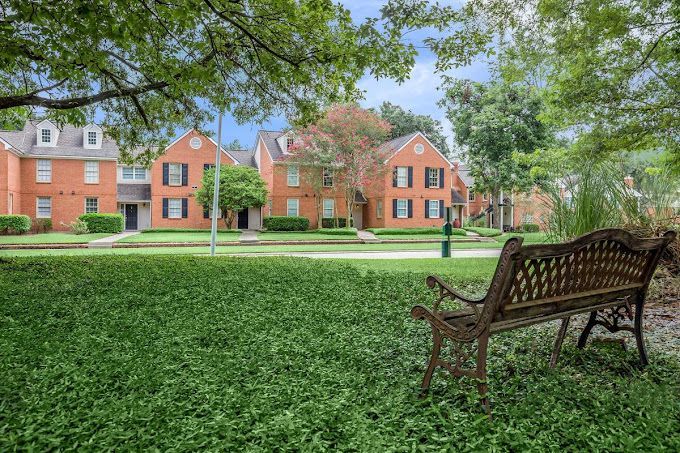 Brick apartment buildings with green lawn and iron bench under a tree.