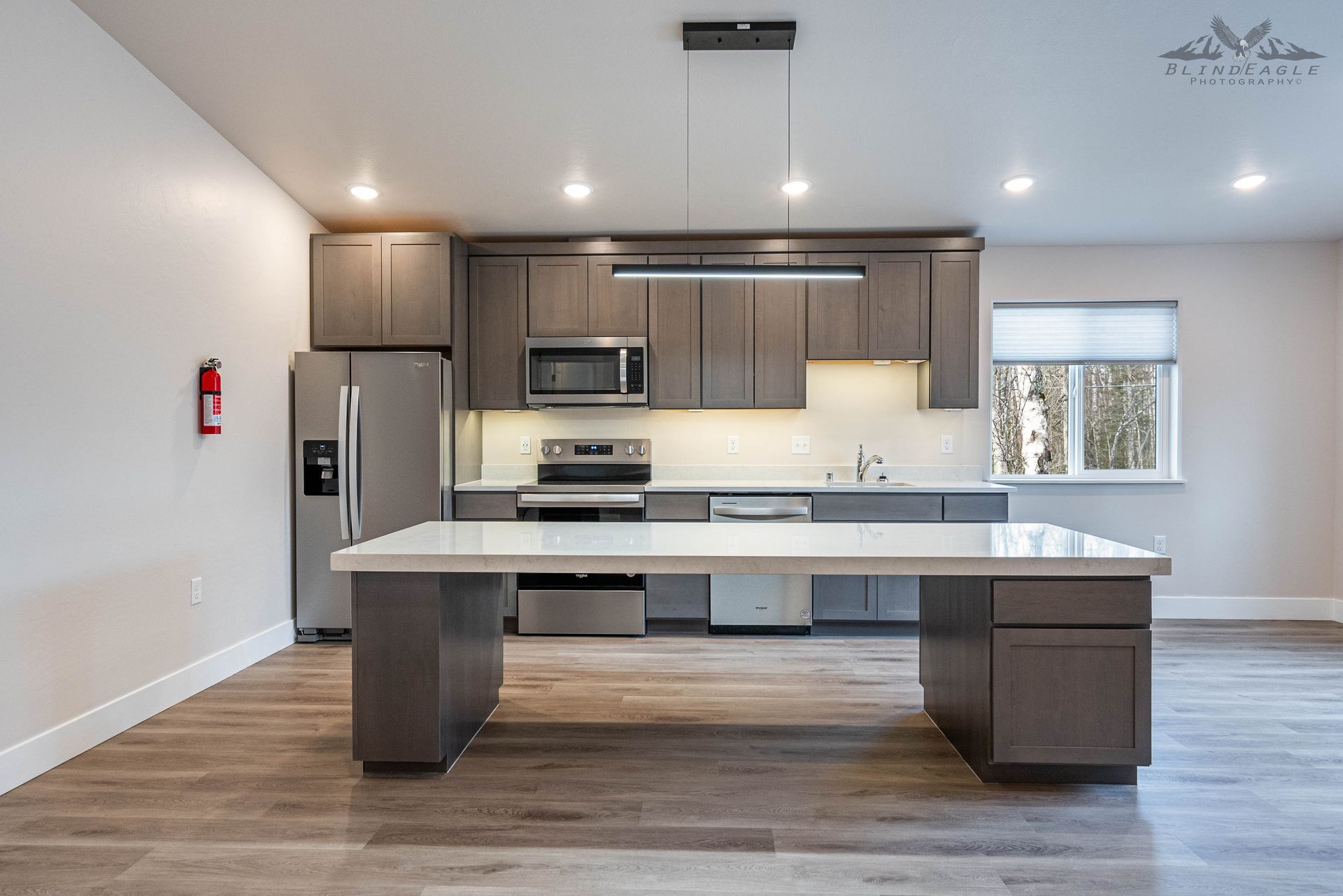 Modern kitchen with a long white-topped island, dark wood cabinets, stainless appliances, and light wood flooring.
