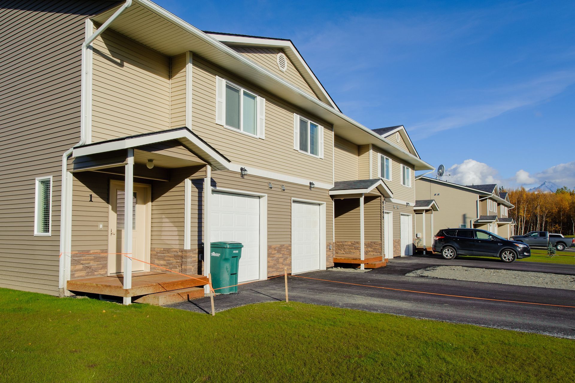 Two-story suburban townhomes with beige siding, attached garages, and a paved driveway under a blue sky.