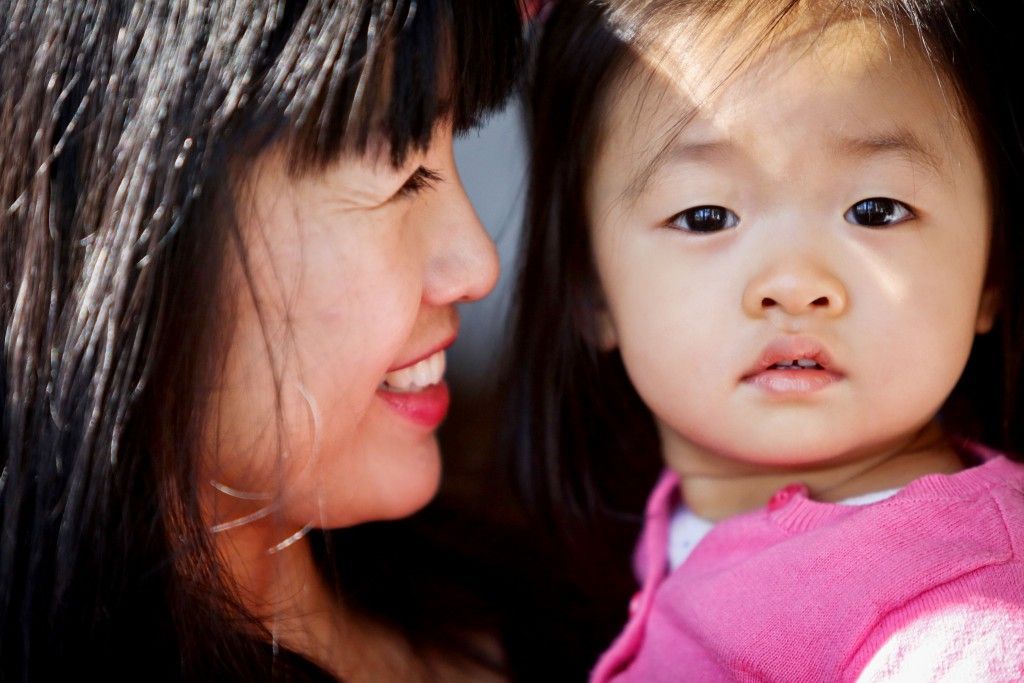 A smiling person in profile looking at a young child in a pink sweater, who is looking directly at the camera.