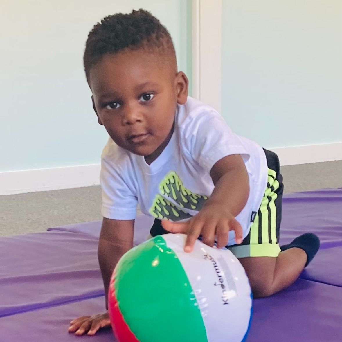 Young child on hands and knees reaching for a colorful beach ball, on a purple mat.