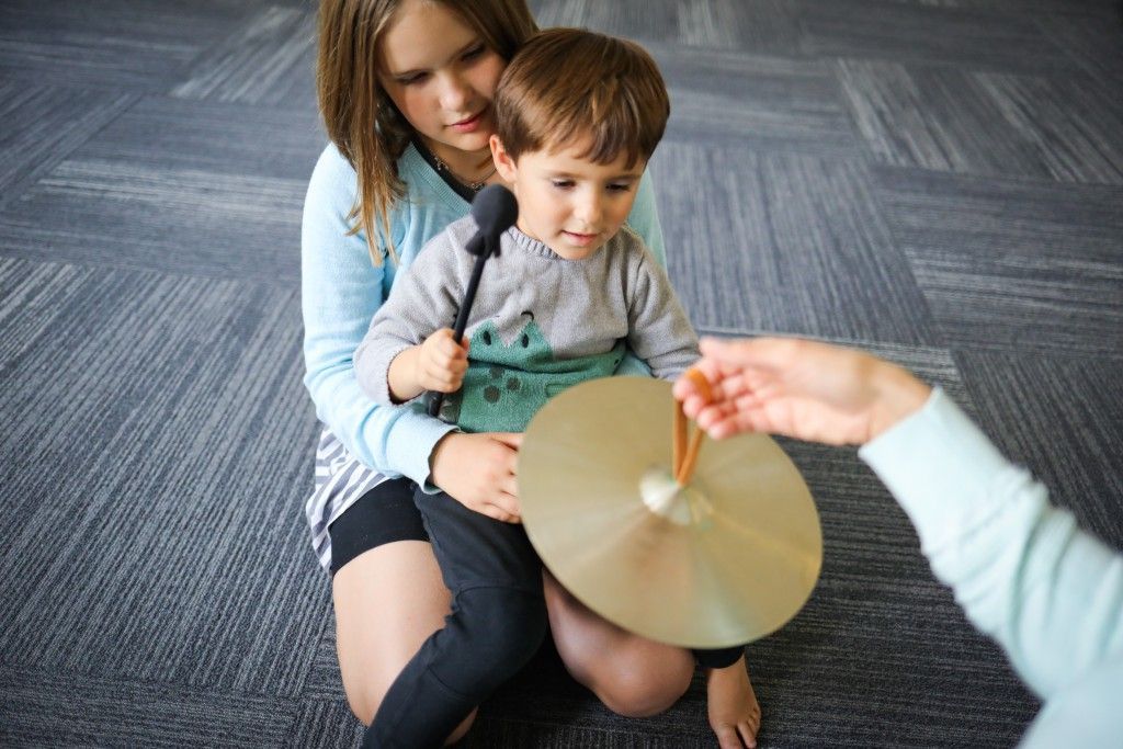 Child playing a cymbal with help. A girl holds the child on her lap, while an adult points to the cymbal. Indoor setting.