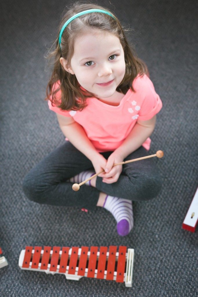 Young girl with a teal headband, holding a mallet, smiling at the camera, sitting on the floor with a xylophone.