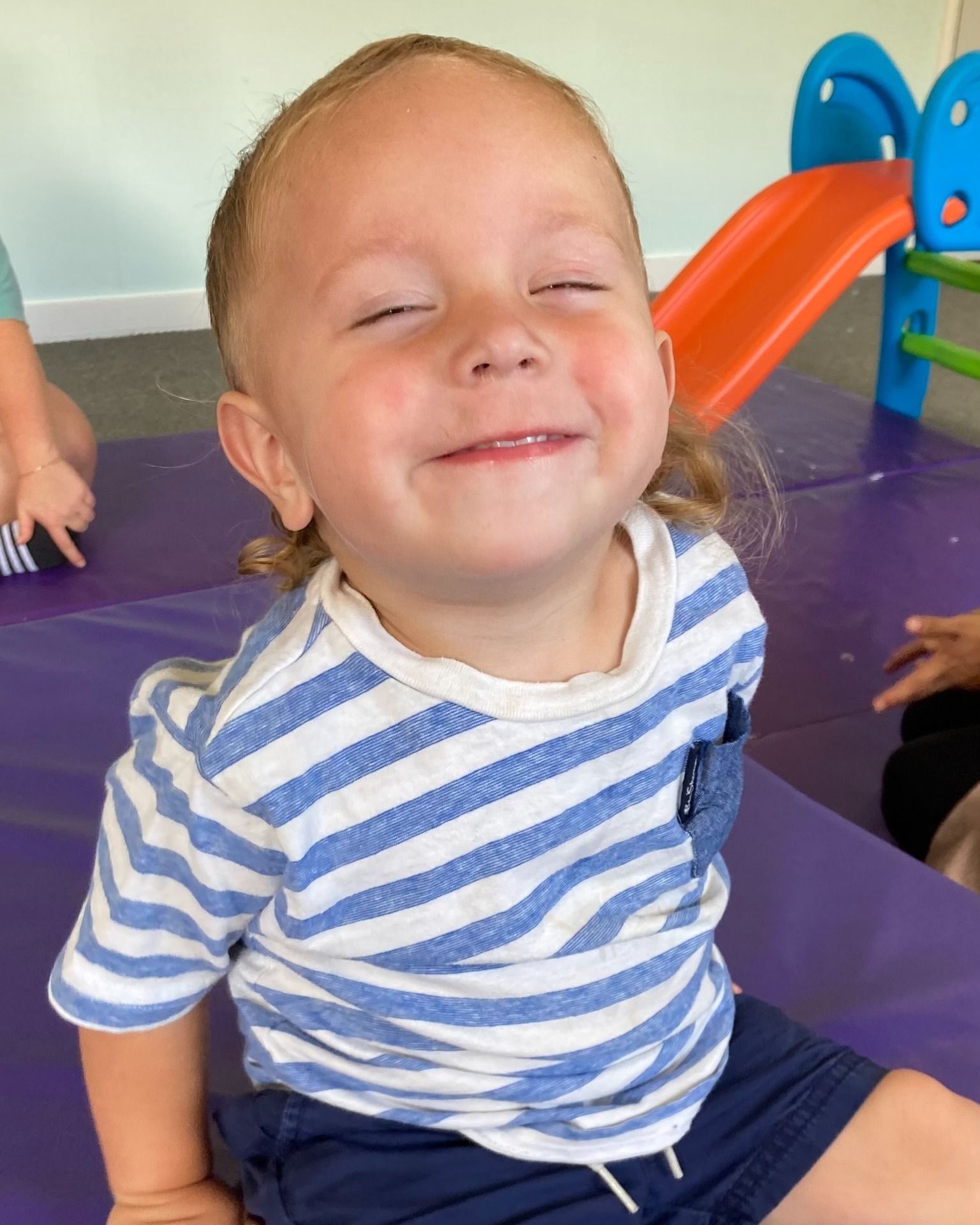 Smiling child wearing striped shirt sitting on purple mat; play area in background.