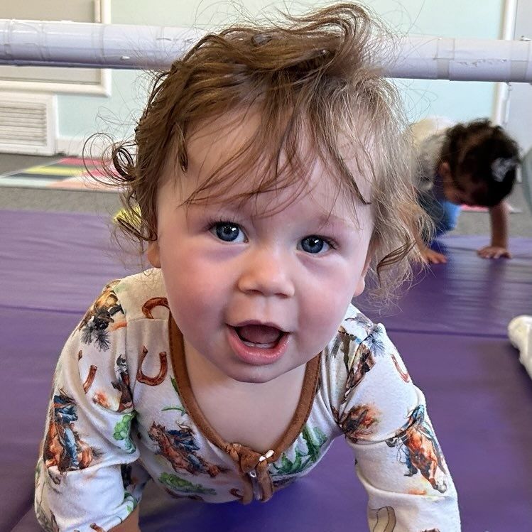 Smiling baby crawling on a purple mat; another child crawls in the background.