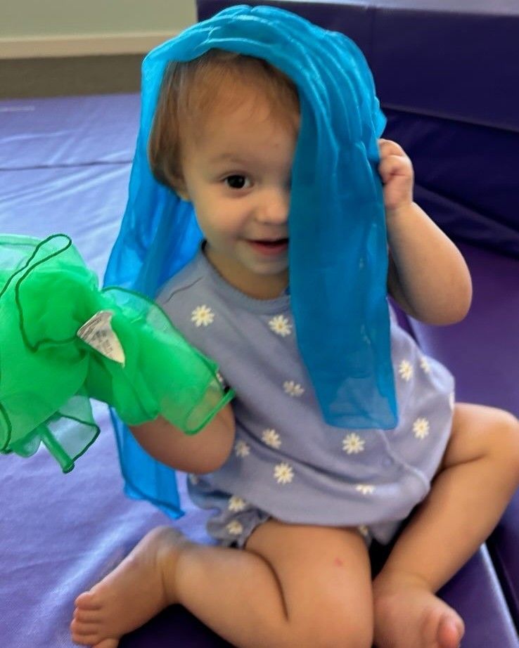 Toddler sitting with blue and green scarves, smiling.