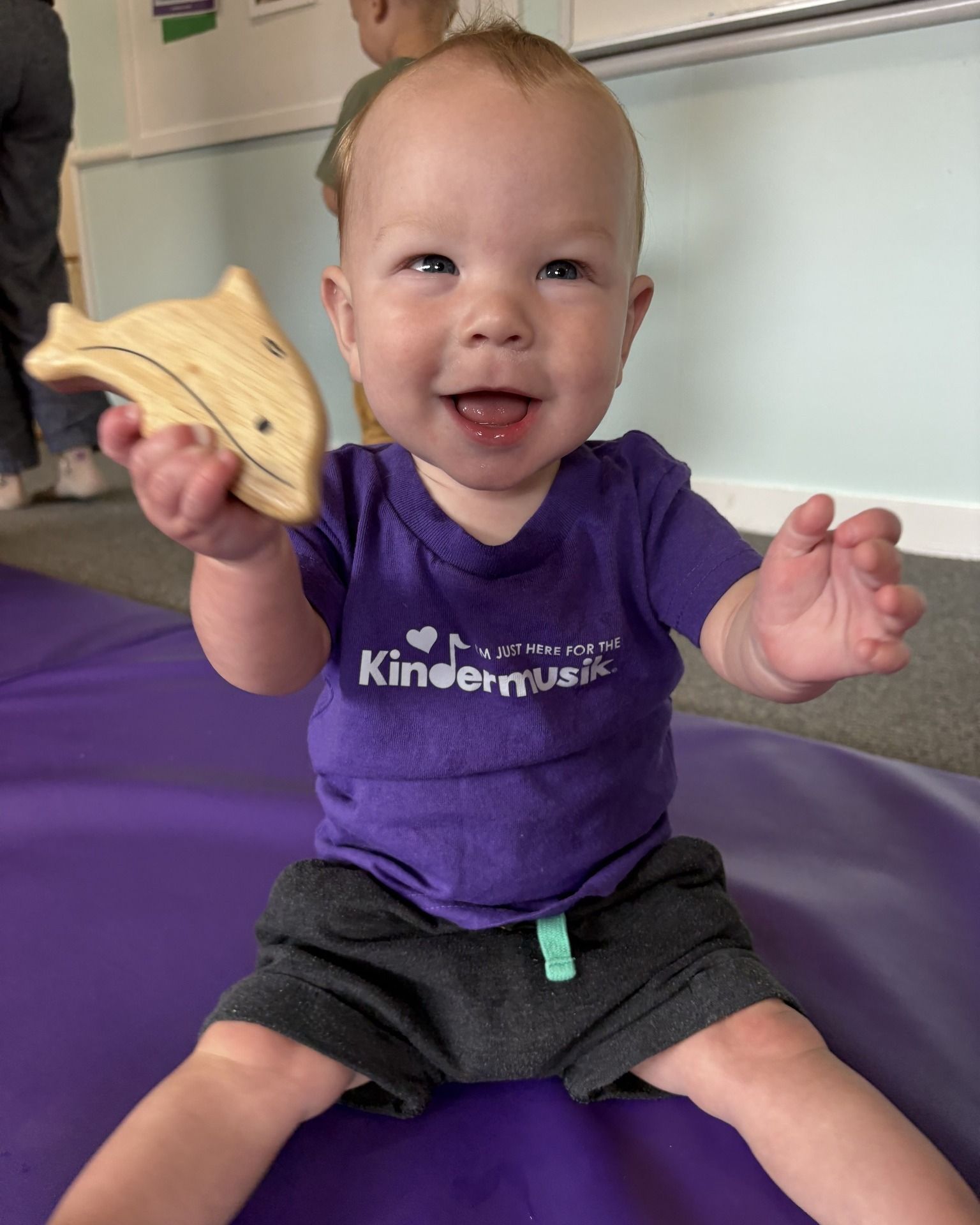 Smiling baby in purple shirt, holding wooden dolphin toy, sitting on purple mat.