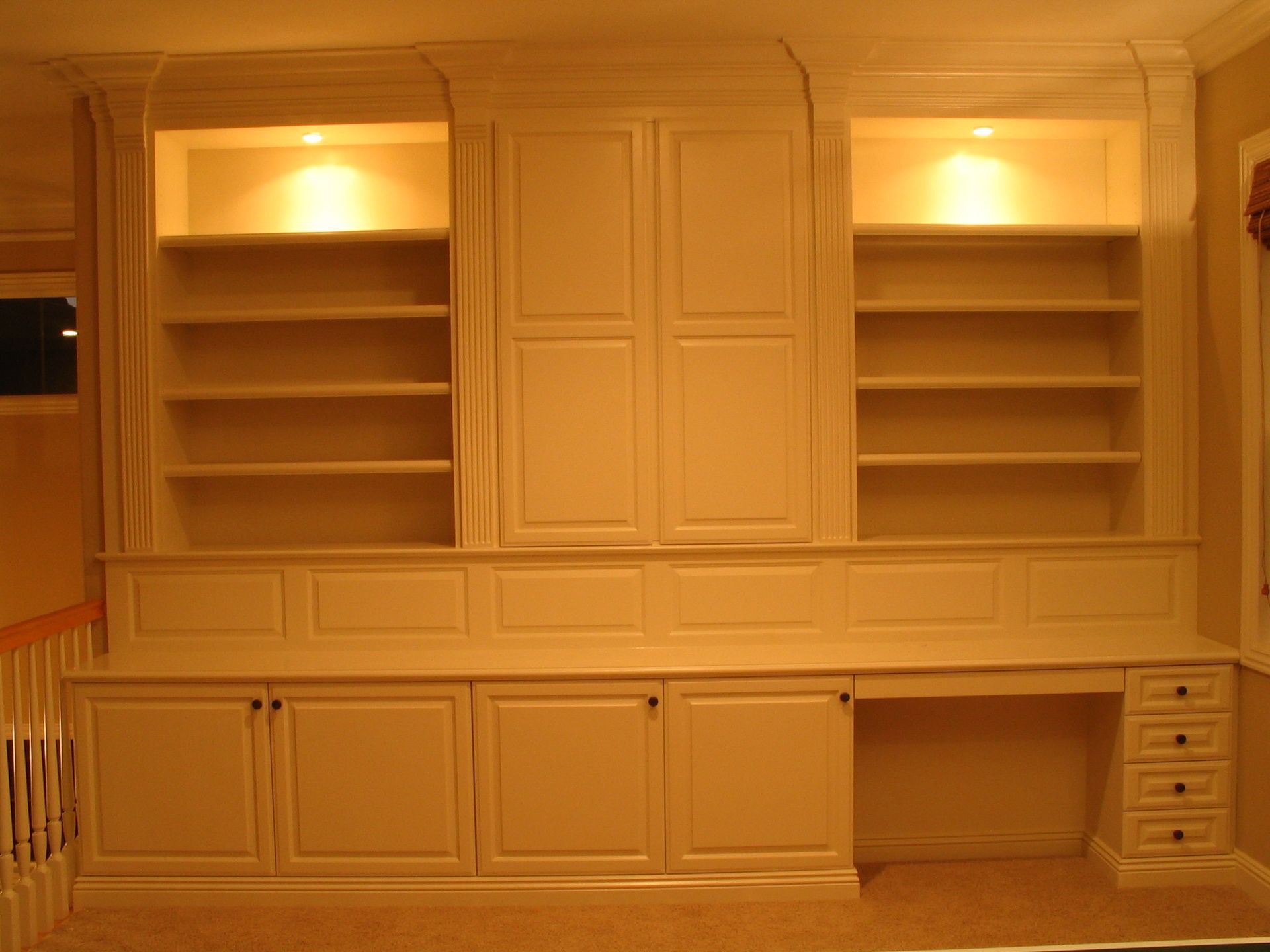 Cream-colored built-in bookcase and desk with overhead lighting. Shelves flank a cabinet, with drawers on the right.