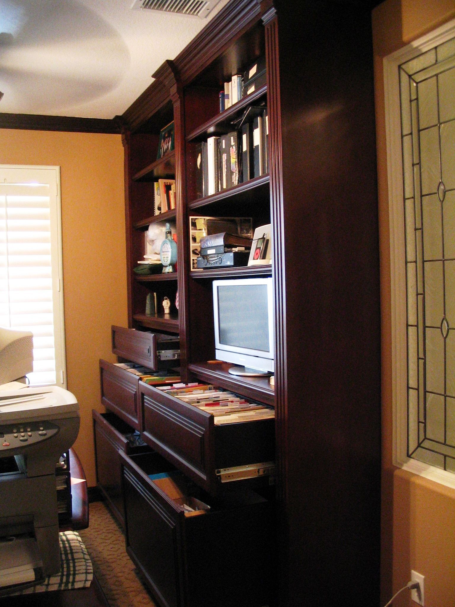 Dark wood built-in bookcase with books, electronics, and drawers, next to a window with decorative glass.