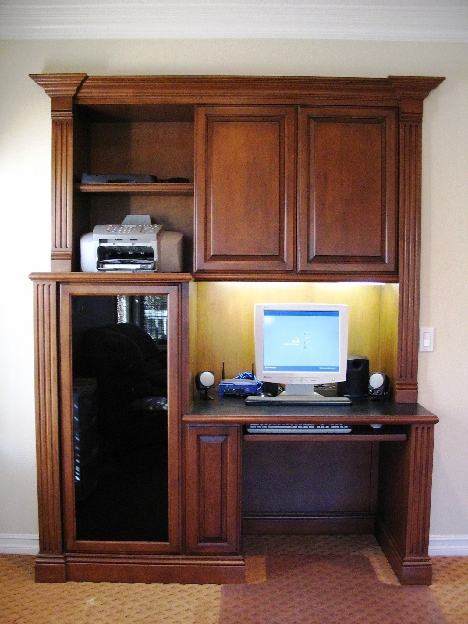 Wooden built-in computer desk with monitor, printer, and cabinets, against a beige wall.