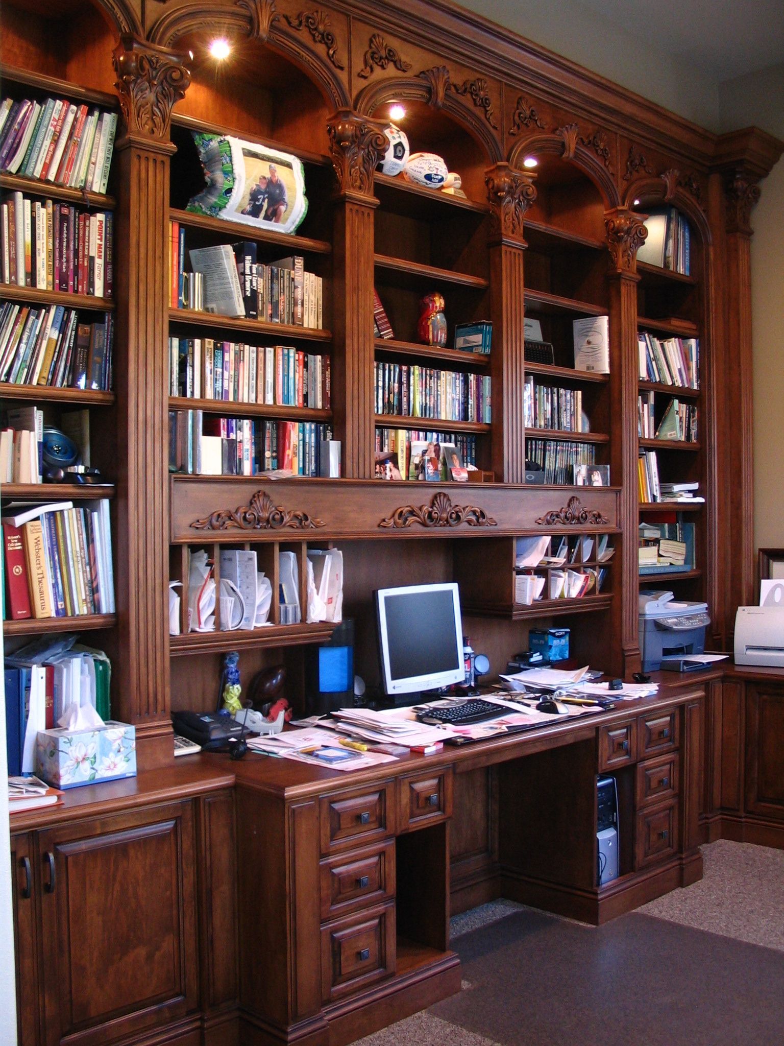Wooden home office with built-in bookshelves, desk, computer, and printer. Dark wood, books, and various office supplies.