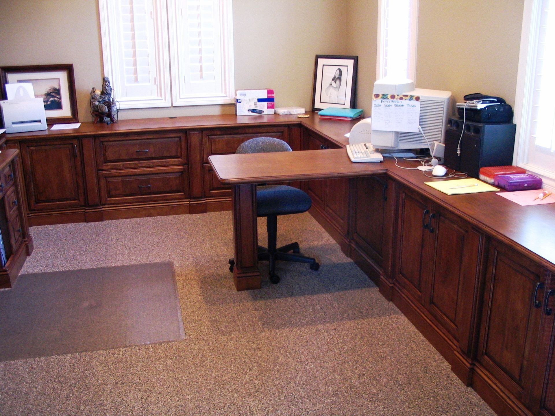 A wooden U-shaped desk in an office with a chair, computer, printer, and window with shutters.