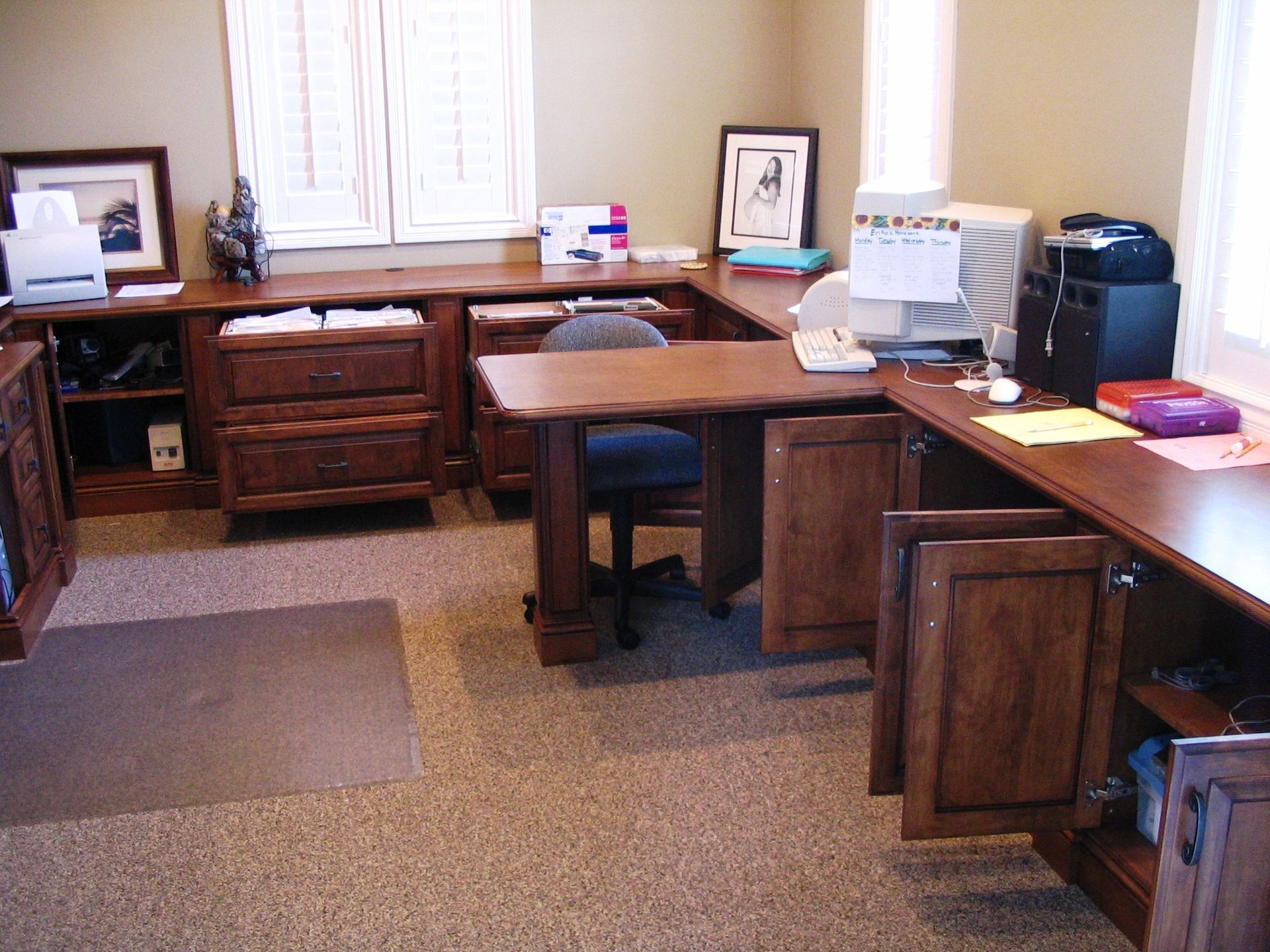U-shaped wooden desk in home office, with cabinets, drawers, computer, and chair. Window and neutral carpet.