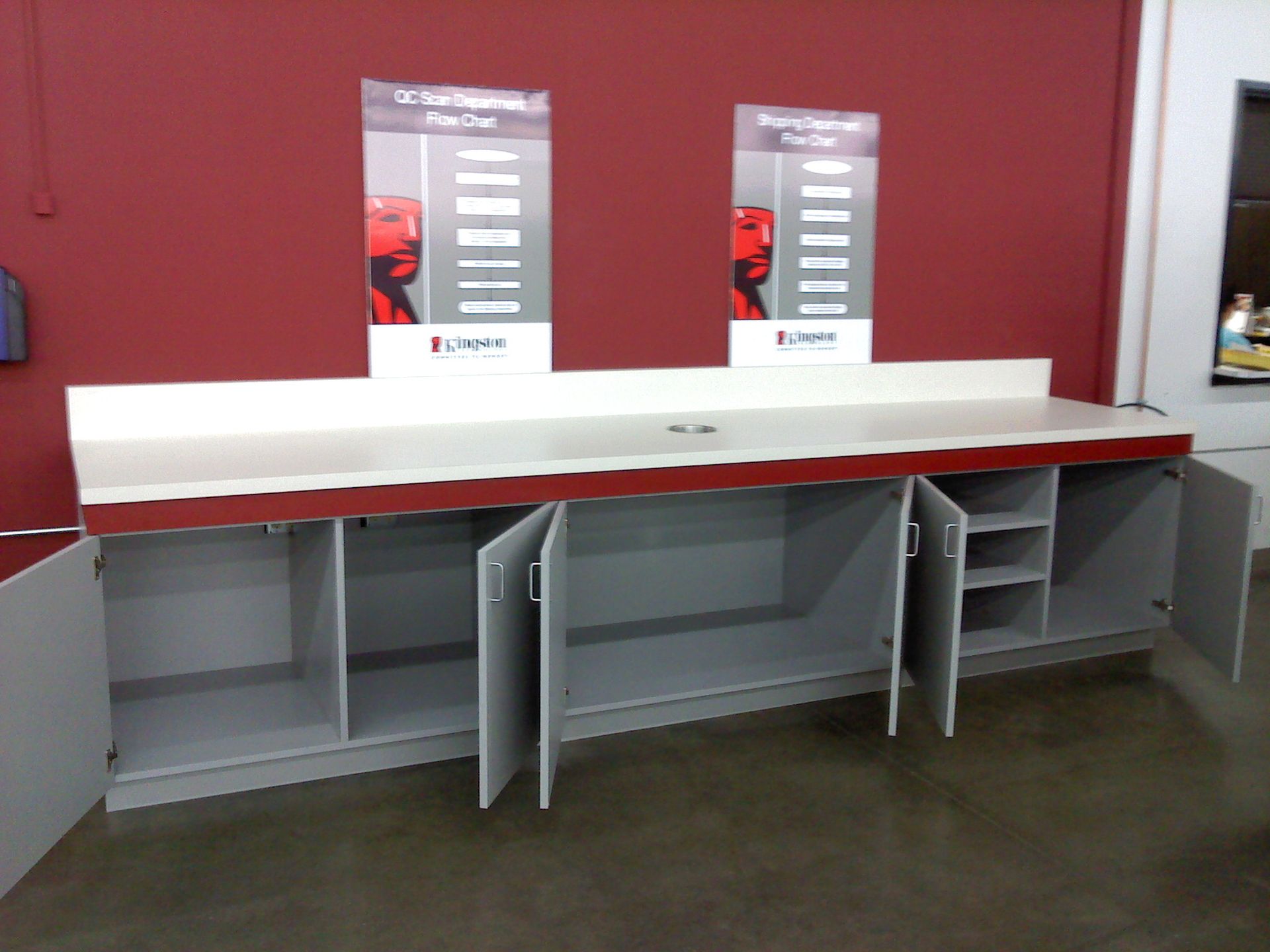 A long cabinet with open doors and a white countertop. Two signs with red accents and a dark red wall are visible.