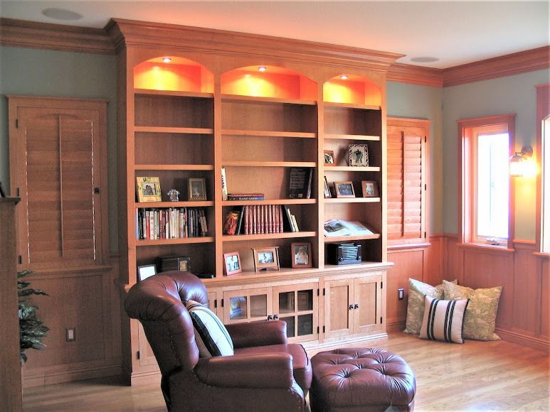 Built-in wooden bookshelf with books and decor, lit from above, in a room with a recliner and ottoman.