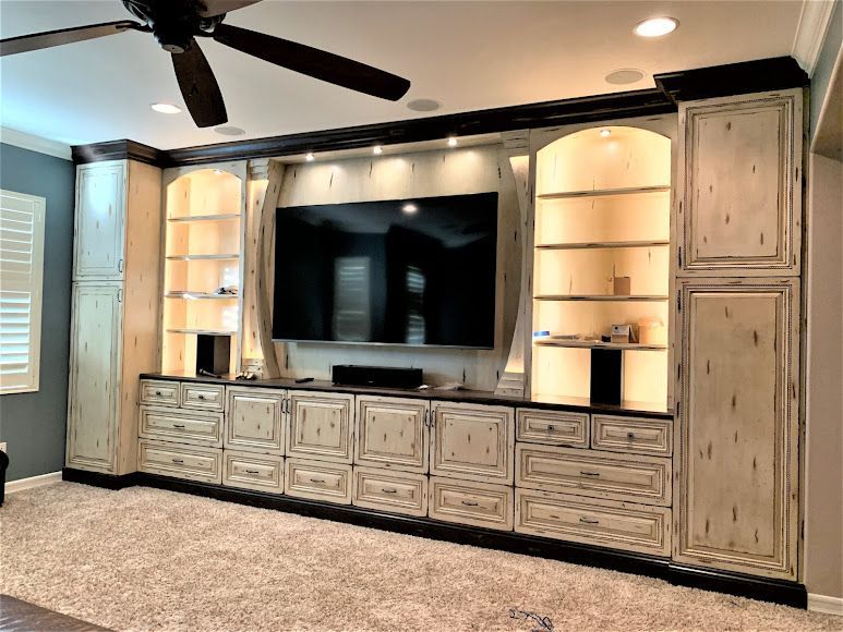 Built-in entertainment center with TV, shelves, and cabinets in a cream-colored, distressed finish. Brown trim