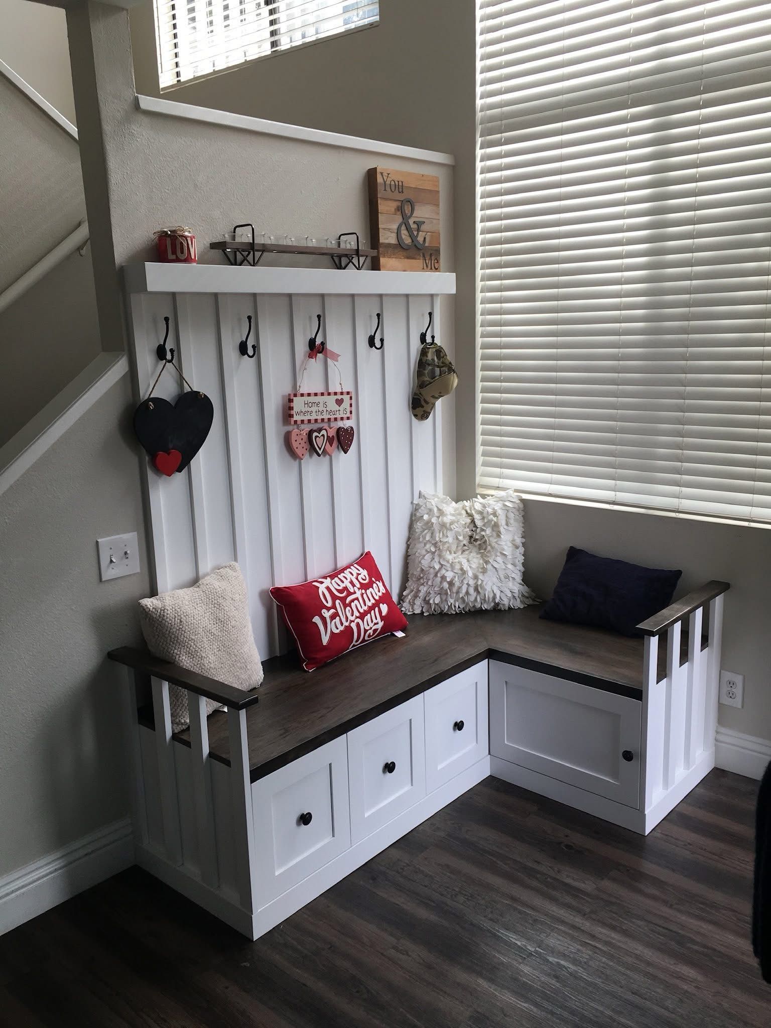 L-shaped white bench with built-in storage and coat hooks. Dark wood top. Pillows and decorative items displayed.