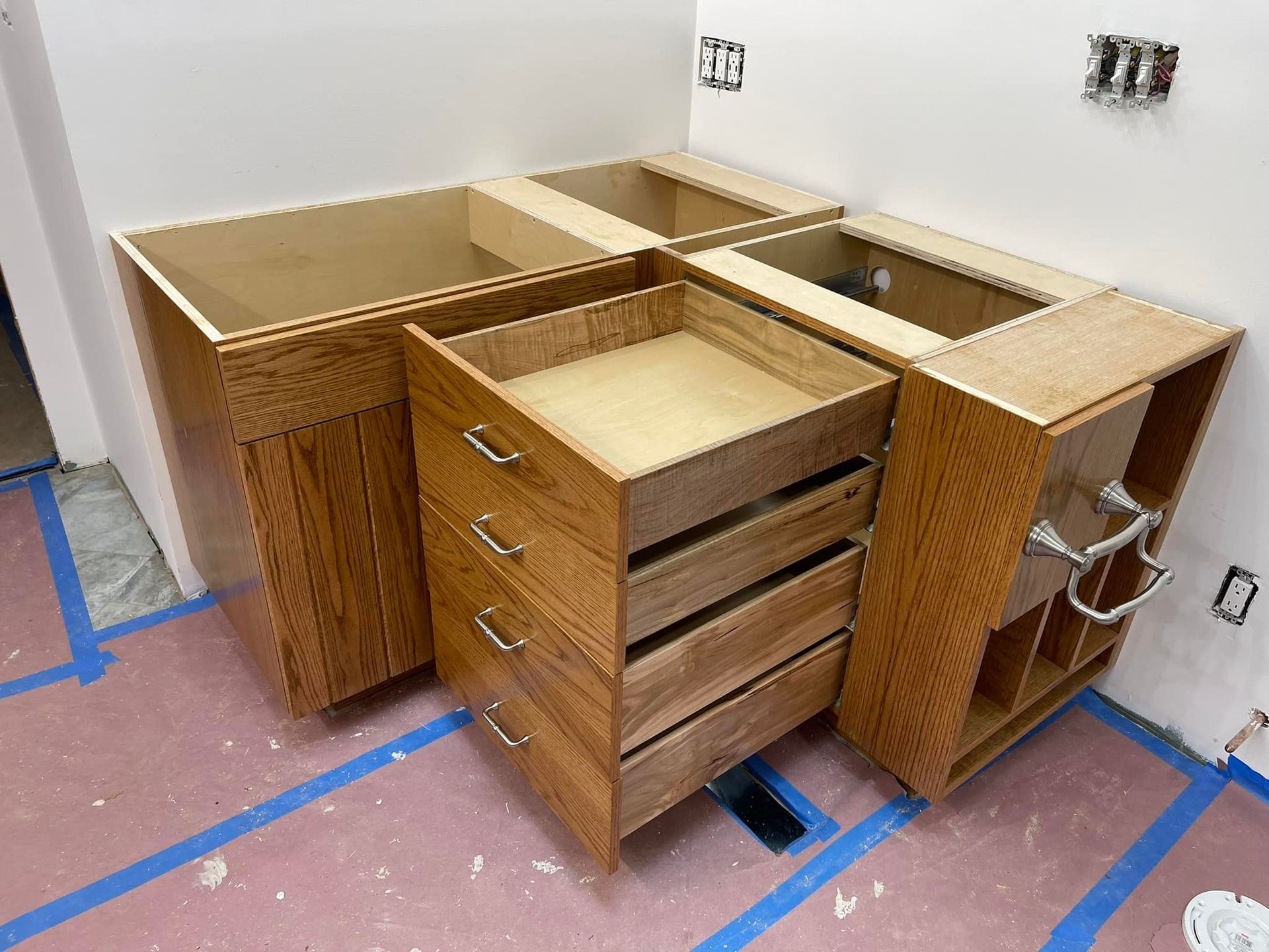 Kitchen cabinets in progress; light wood, drawer unit, open compartments, against white walls, blue tape on floor.