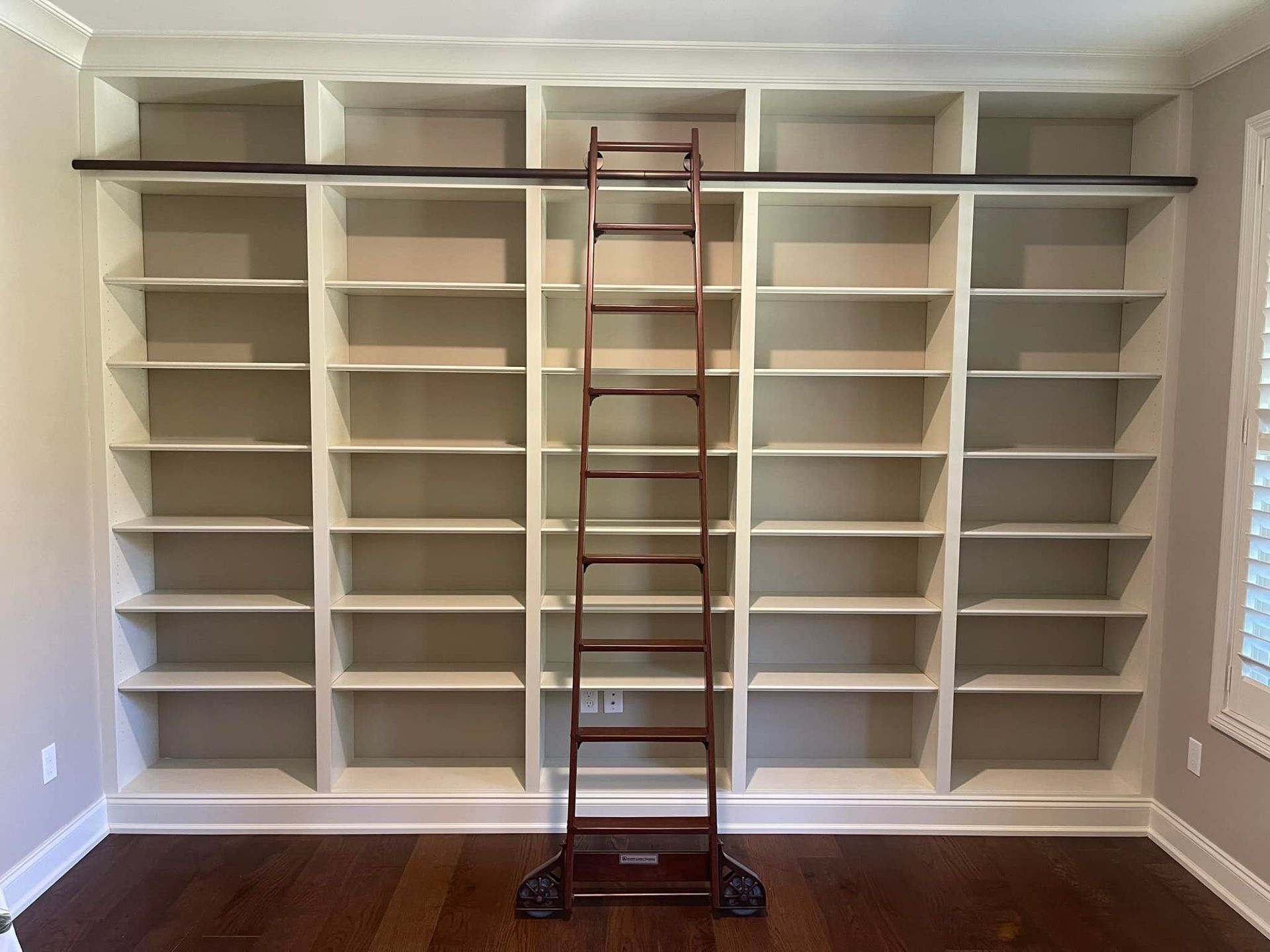 Empty built-in white bookshelves with rolling ladder and black track. Wooden floor and cream-colored walls.