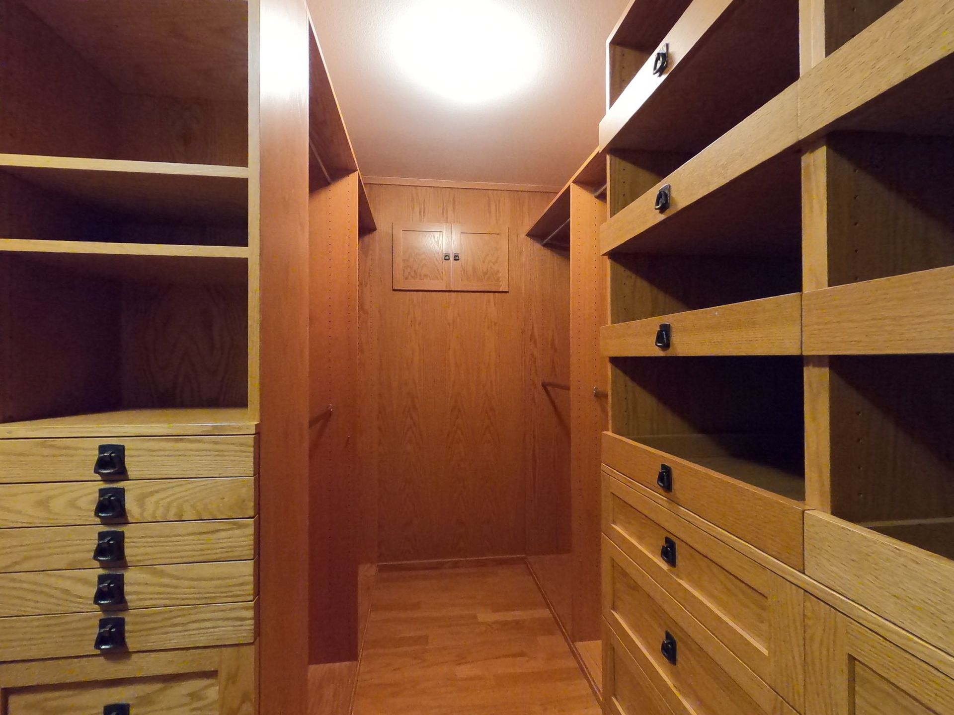 Wooden walk-in closet with shelves and drawers. Soft overhead lighting, wood flooring.