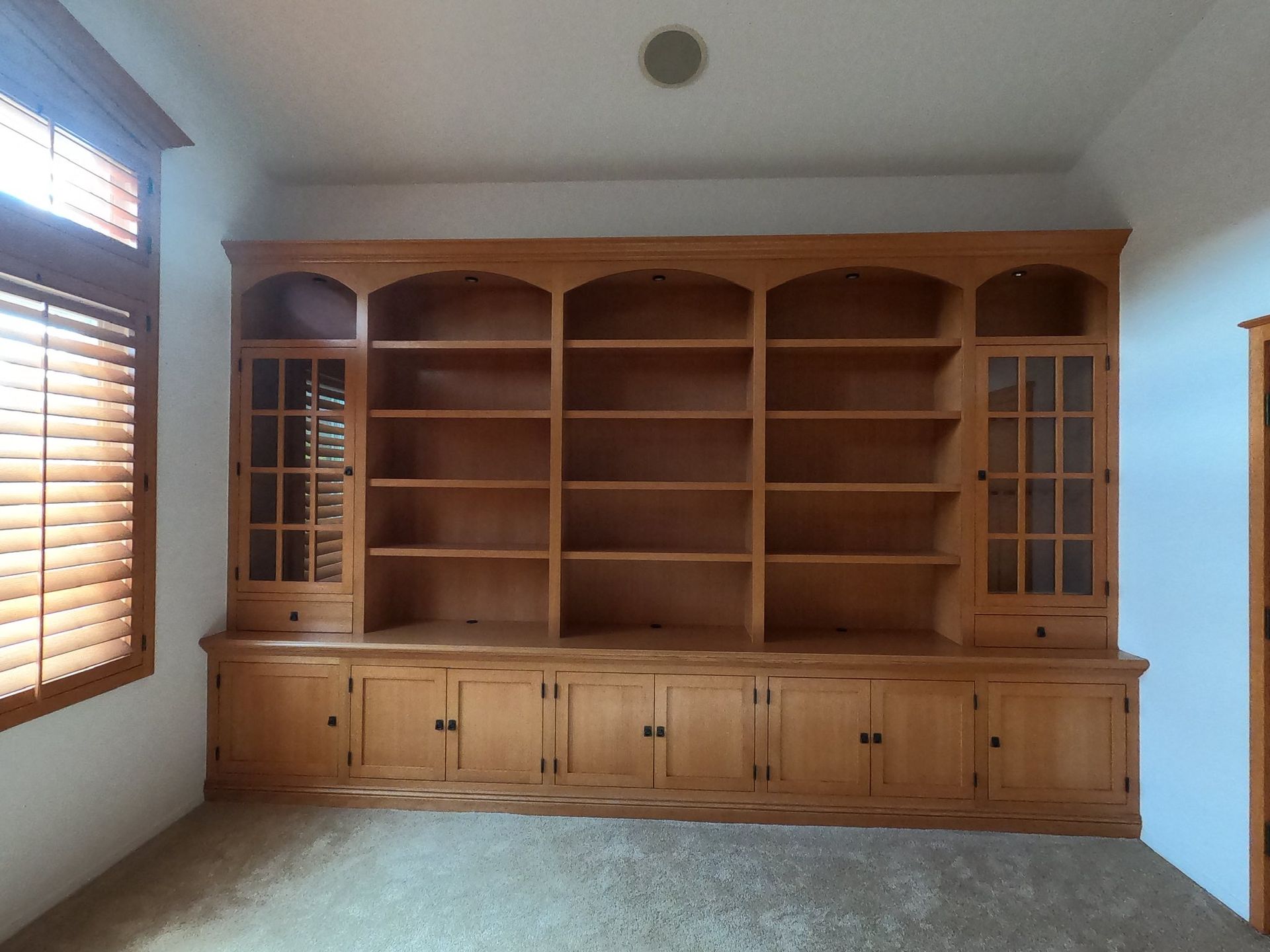 Wooden built-in bookcase with arched shelves, cabinets, and glass-door sections. Light-colored wood in a room with a window.