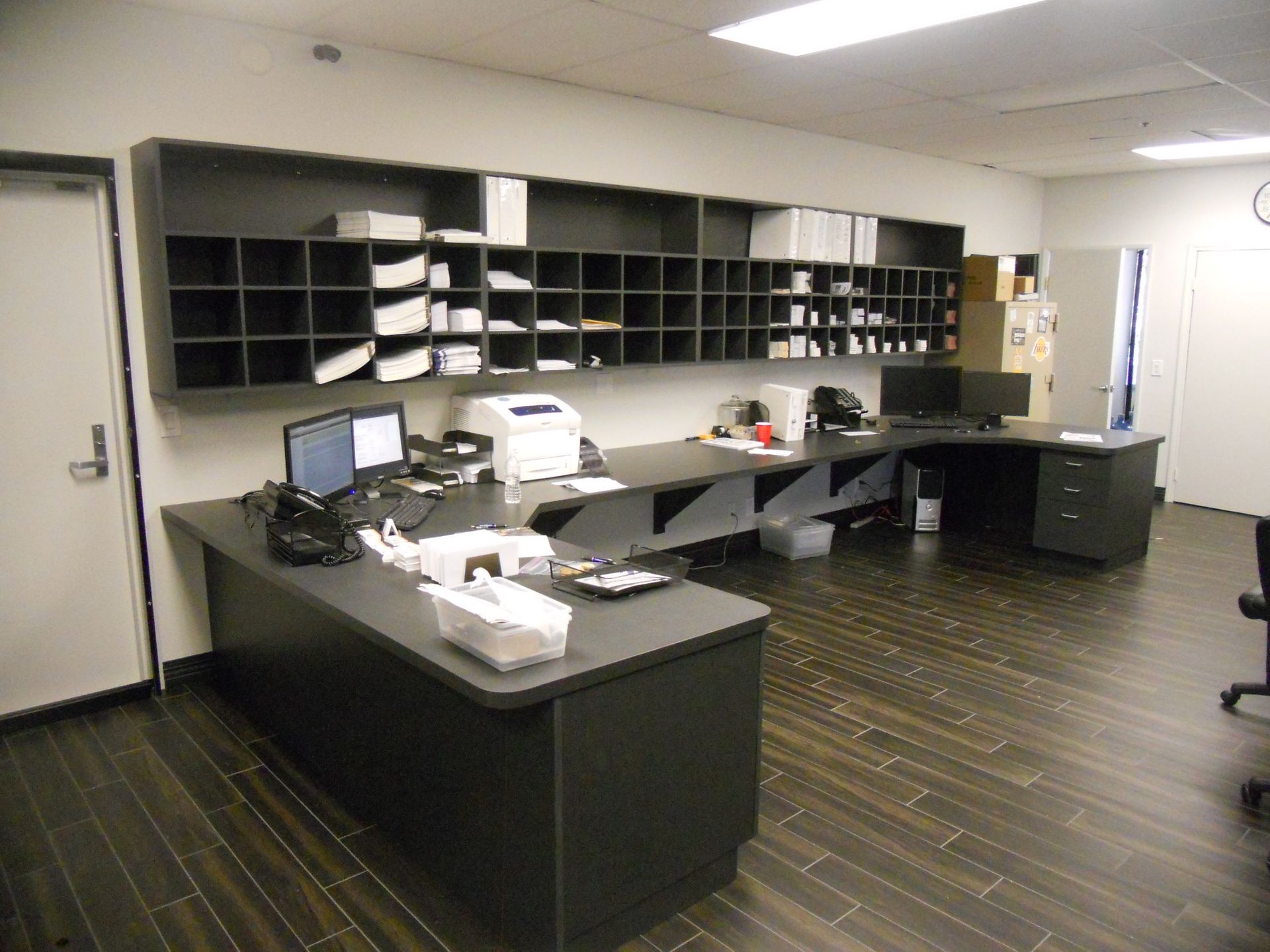 Office workspace with dark desks, mail cubbies, and a printer. Wooden floor.