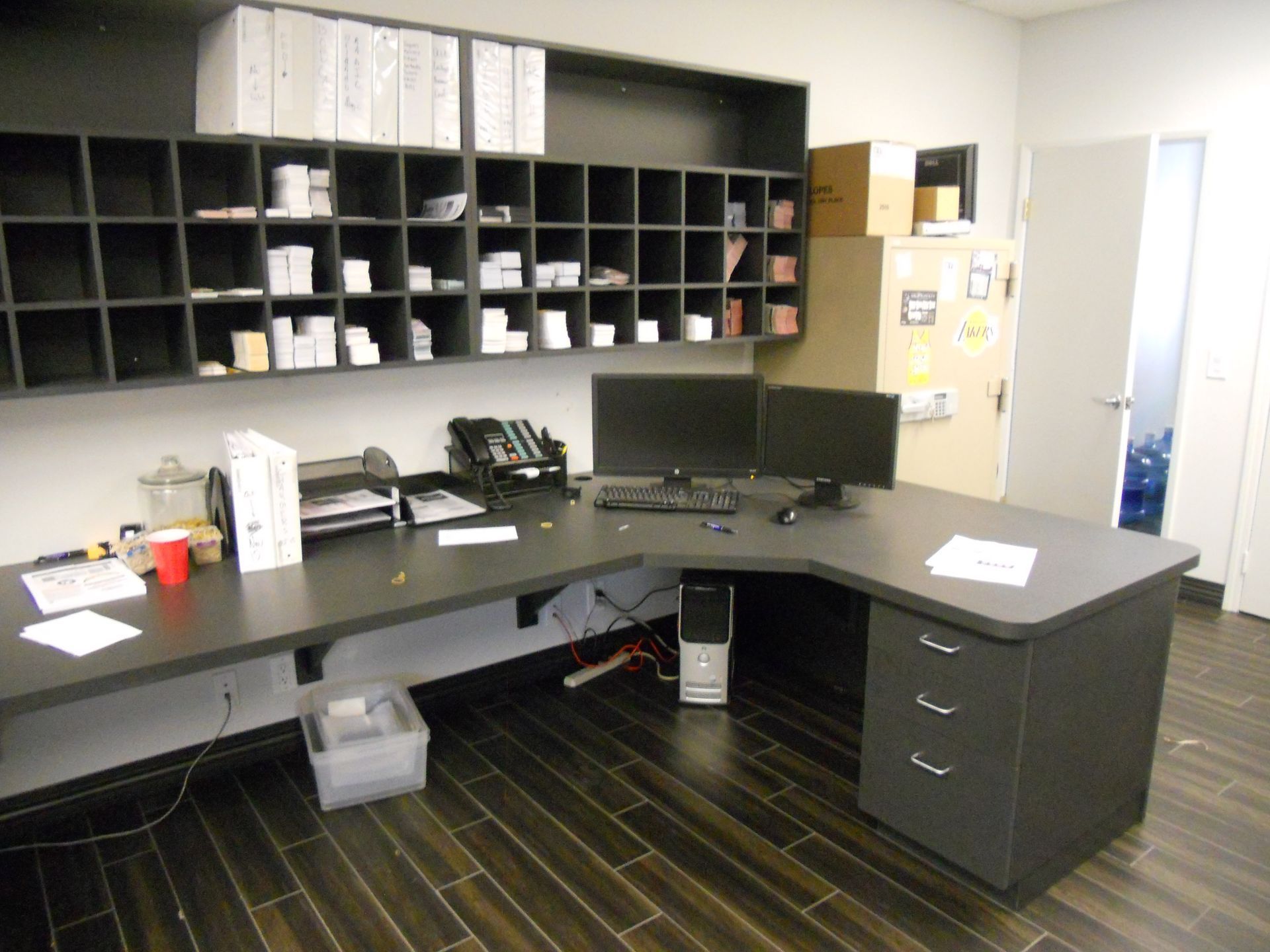 L-shaped office desk with two monitors and storage, shelves above, with a neutral toned color scheme and hardwood floors.