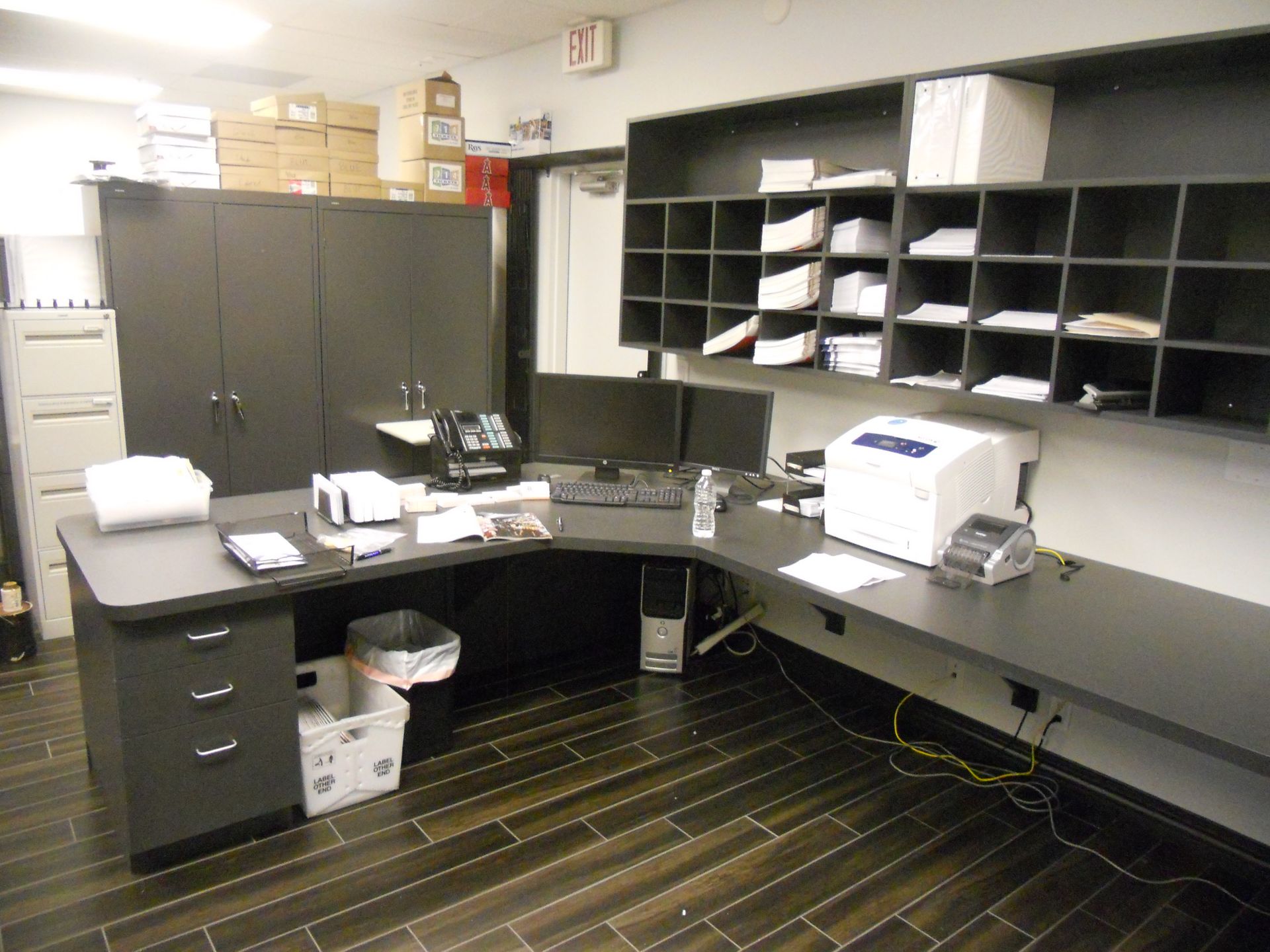 Office space with dark desk, shelves, and storage, featuring a computer setup and mail sorters.