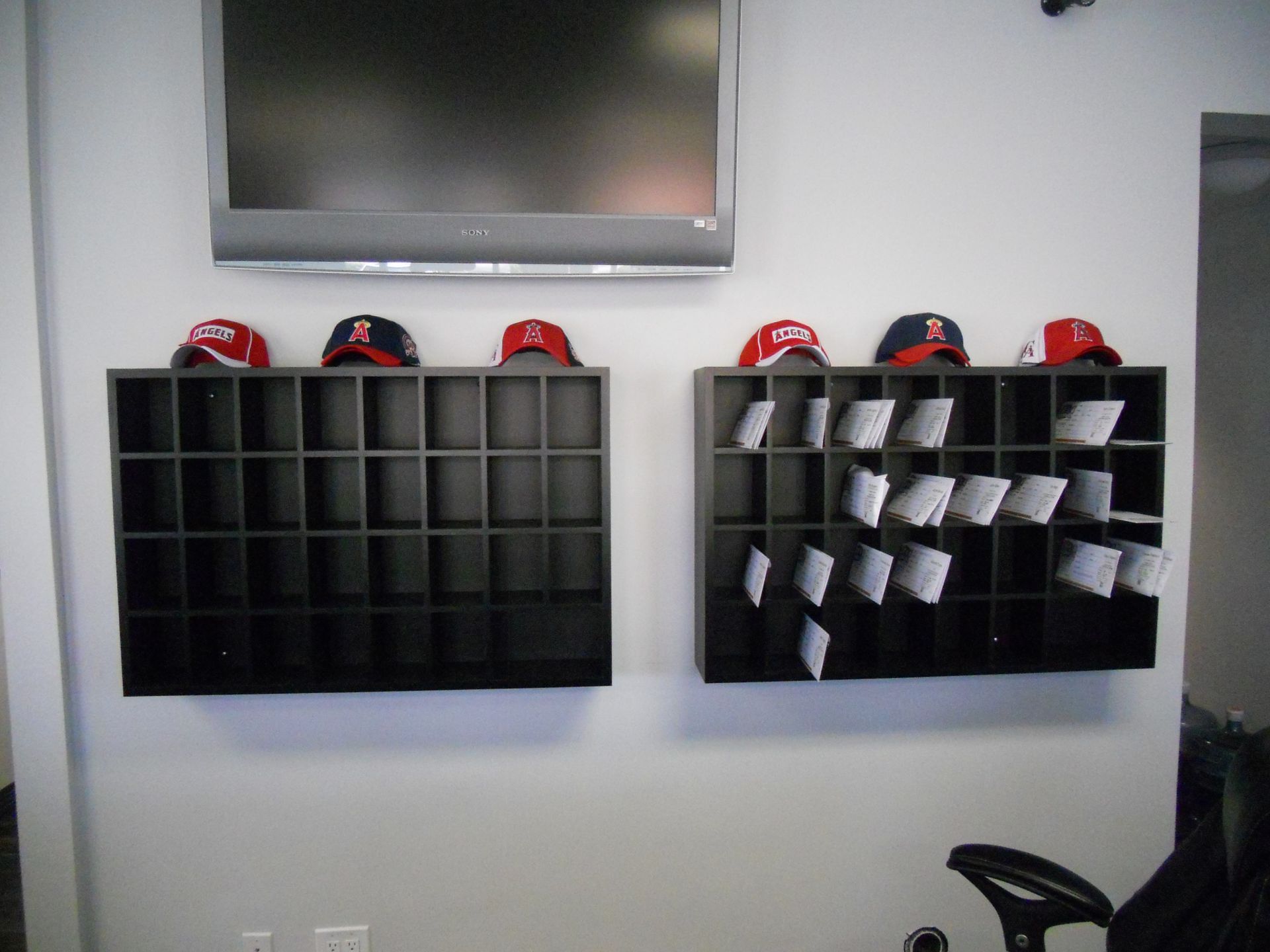 Two black cubby shelves on a white wall; one empty, one with papers, baseball caps above a TV.
