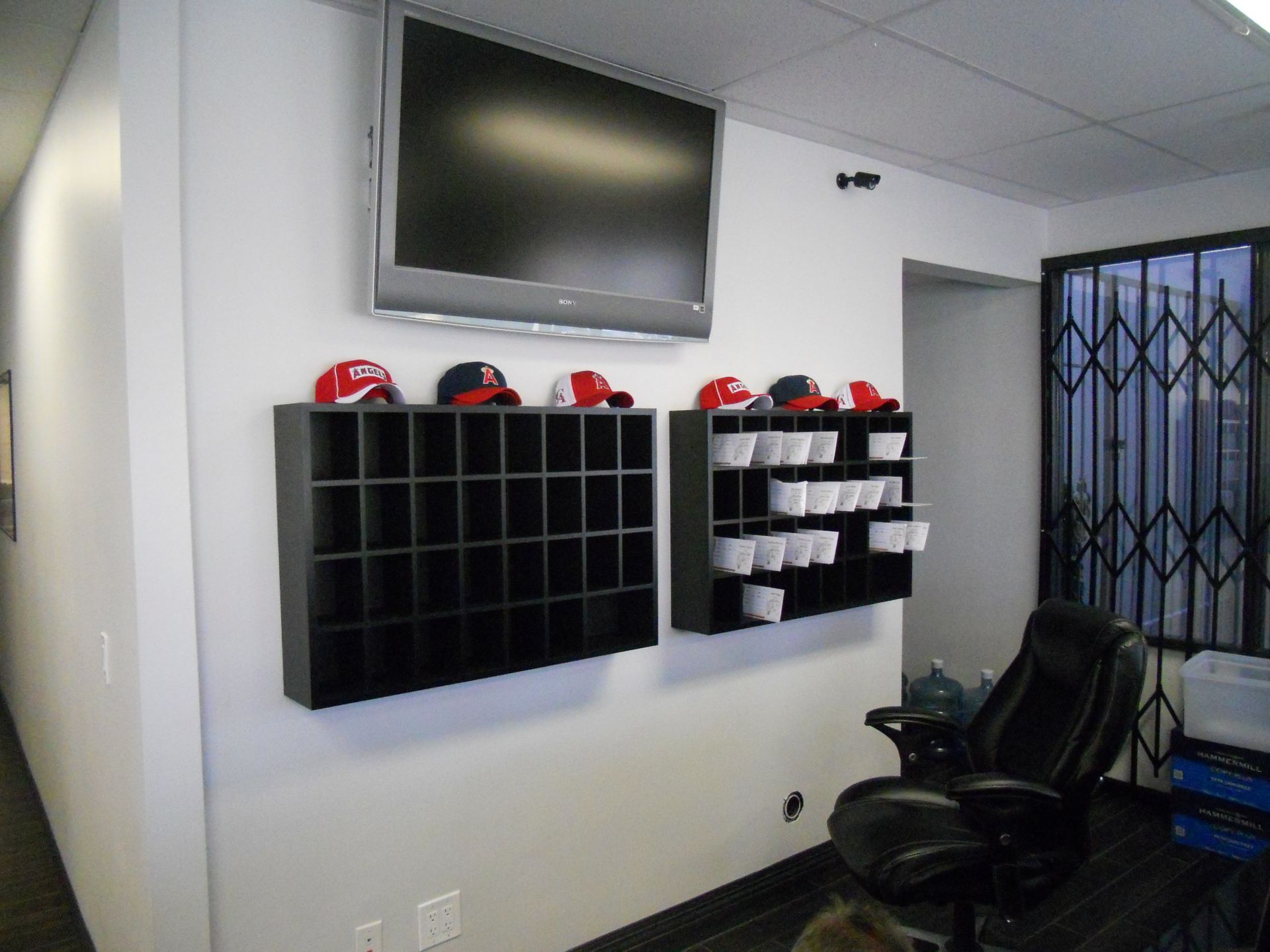 White wall with mounted TV, cubby shelves, baseball caps, and a black chair.