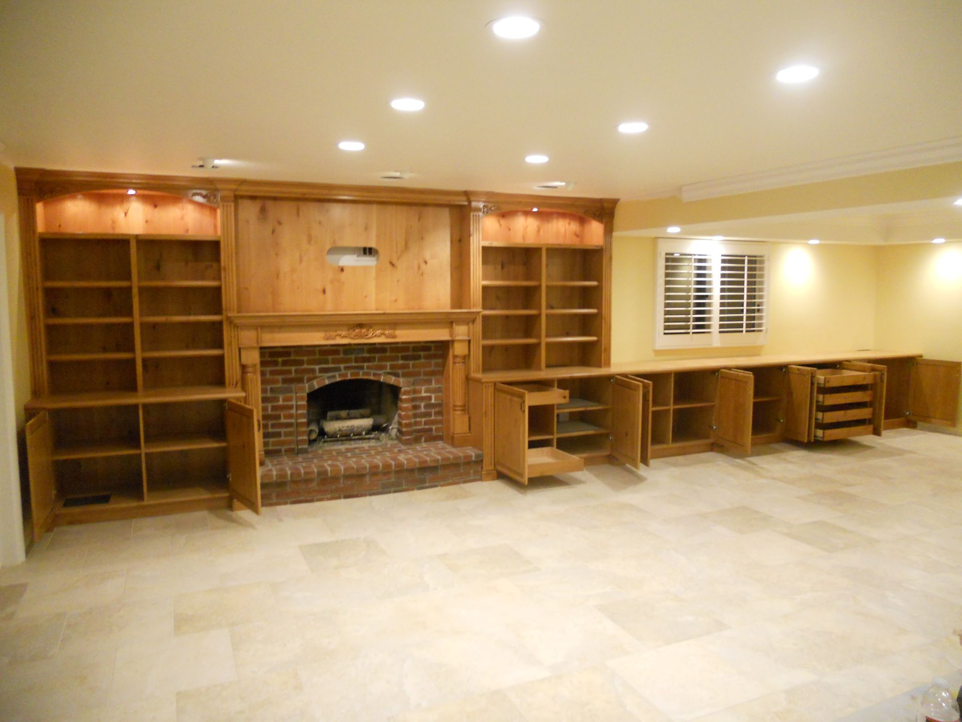 Built-in wooden bookshelves, fireplace, and cabinets in a room with recessed lighting and tiled floor.
