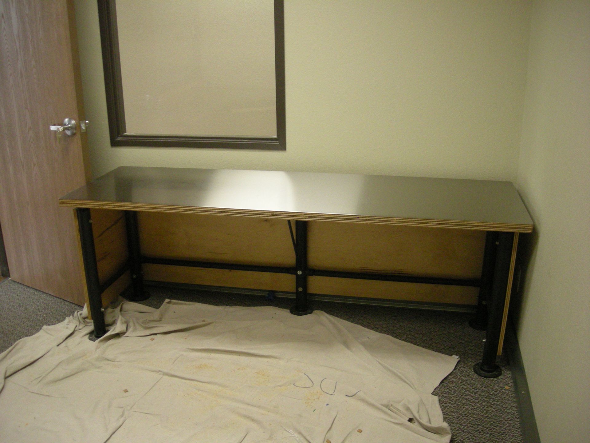 A long, silver-topped desk with black legs, against a beige wall, under a framed mirror.