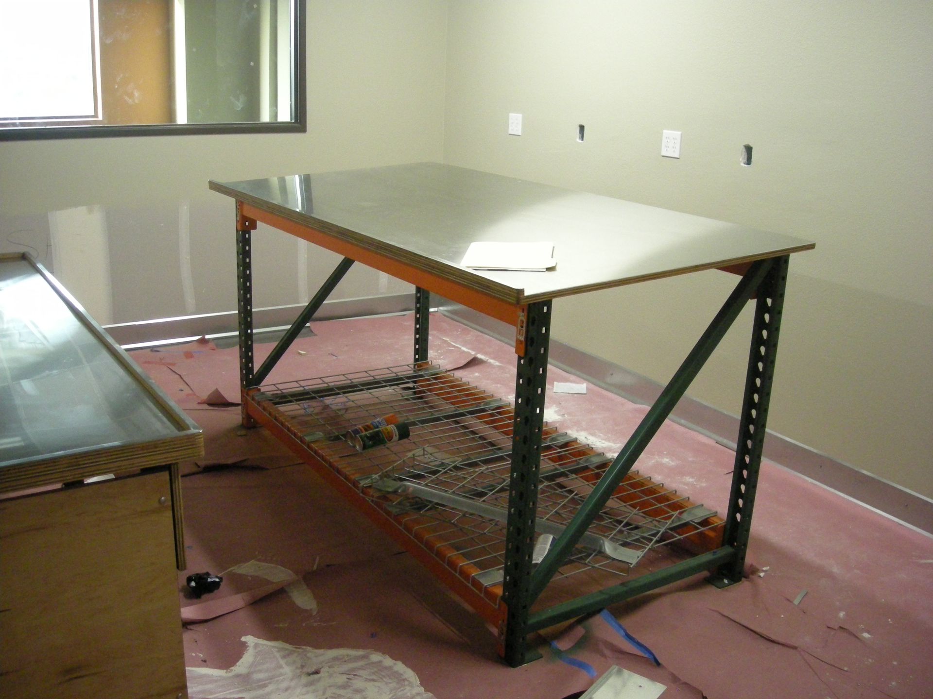 Metal-framed workbench with a metal top and shelf in a room with a window, against a beige wall.