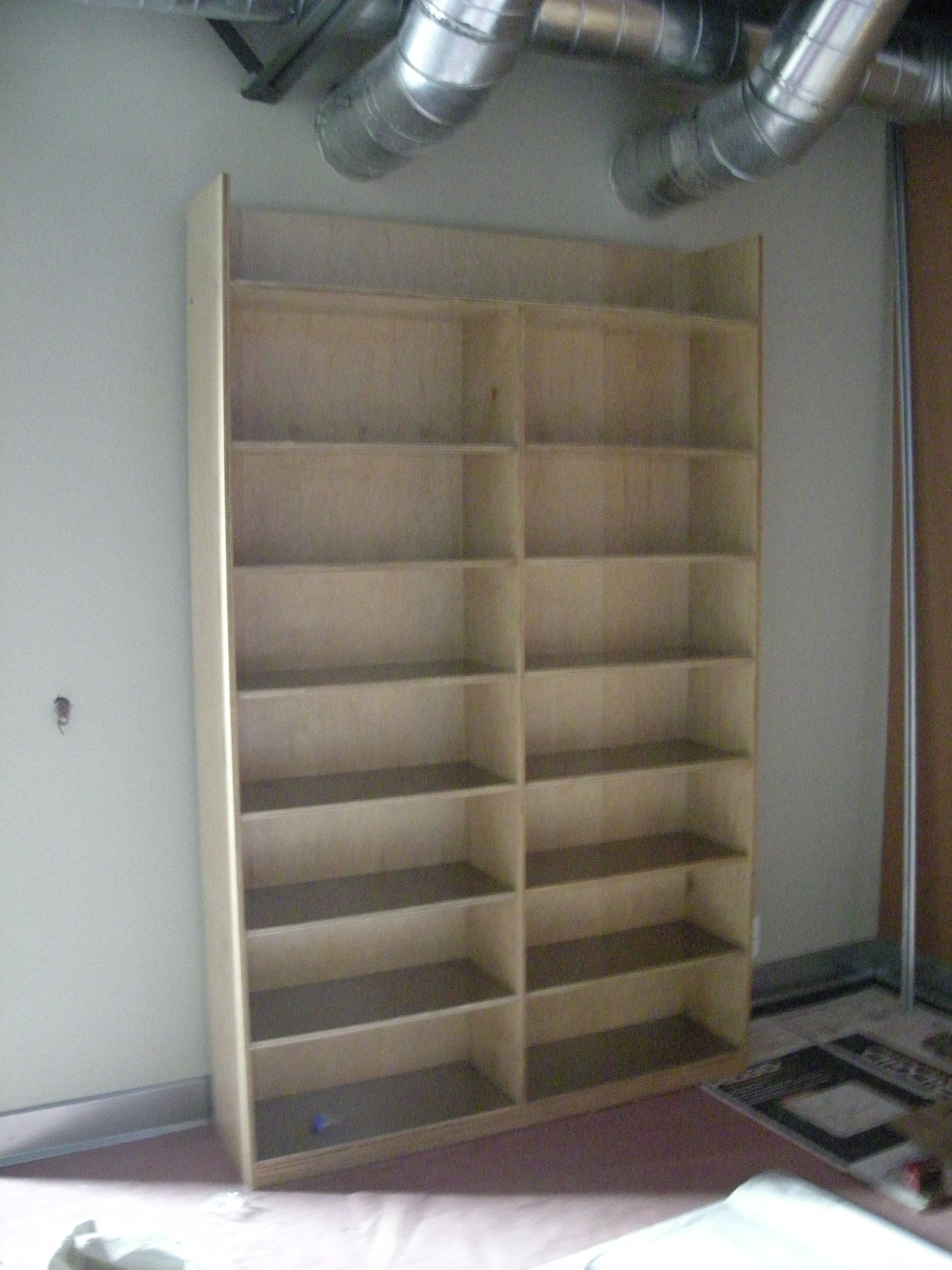 Empty wooden bookshelf against a wall with ductwork overhead.