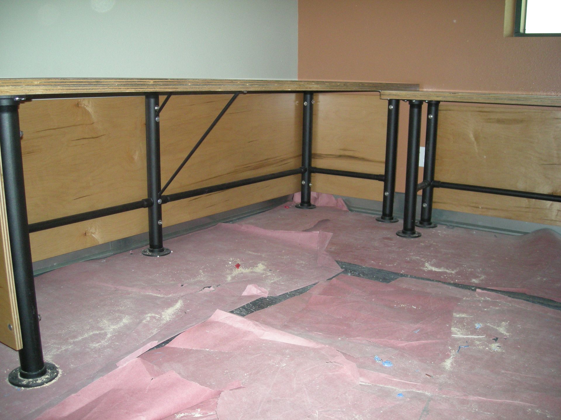 Black metal support posts holding up a wood countertop in a room; floor covered in pink paper.