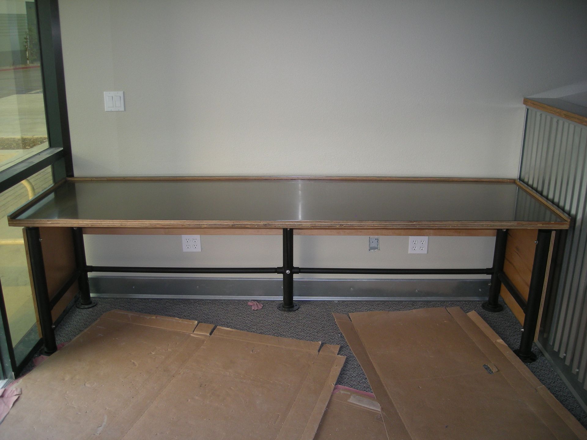 Long rectangular desk with metal top and wood and black metal legs against a beige wall.