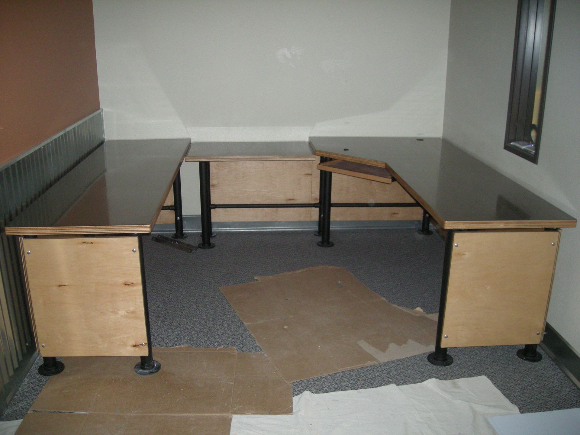 U-shaped desk with a dark top, light wood sides, and black legs, in a room with neutral walls.