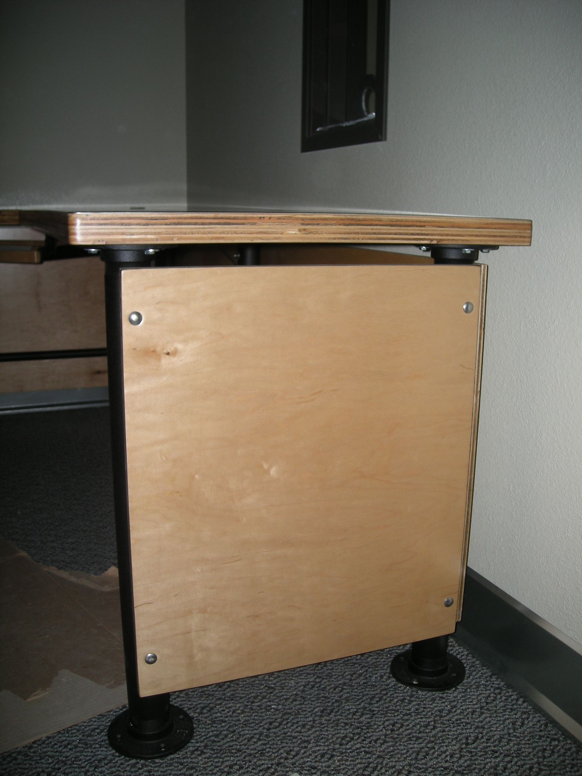 Wooden desk corner with a light-colored side panel, black supports, and adjustable feet.