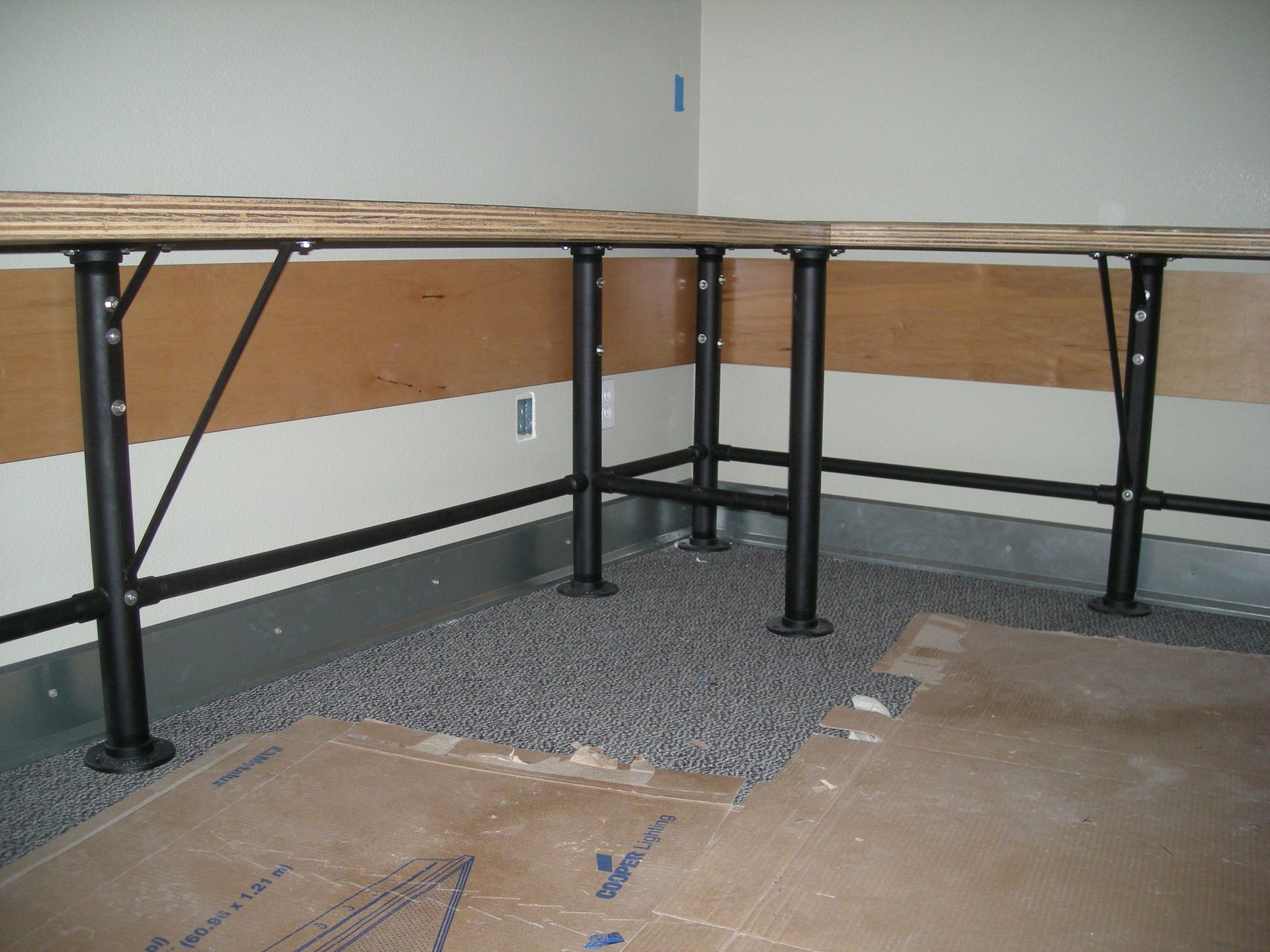 Black metal table supports on gravel-covered floor with plywood tabletop against a wall.