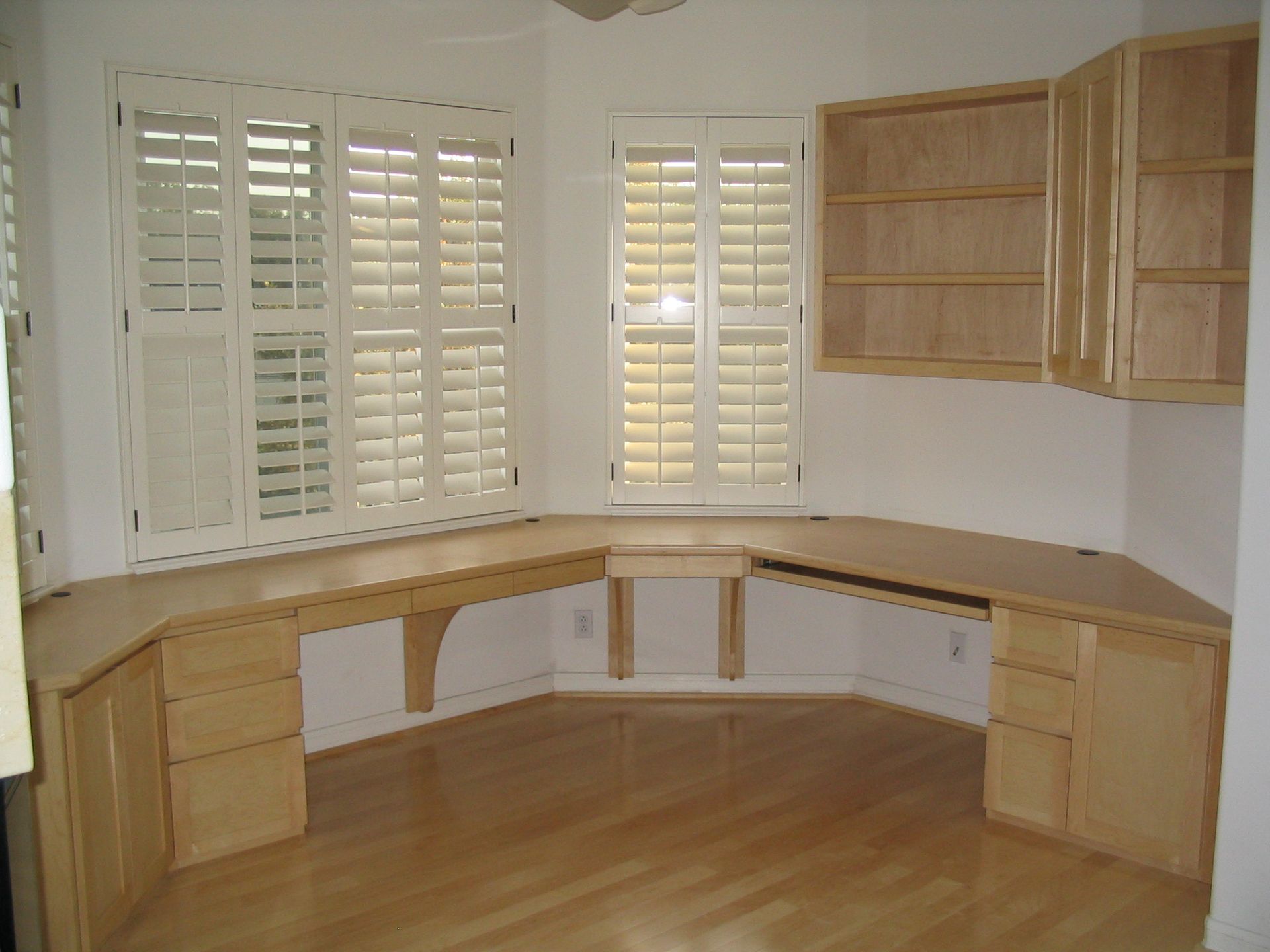 Corner office with built-in light wood desk, cabinets, and two shuttered windows. Hardwood floor.